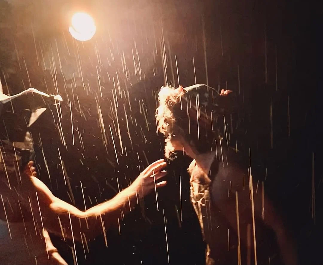 A person with animal ears and a nose, possibly a theatrical or costume character, is touching another person's face under rain illuminated by a bright light overhead in a dark setting.