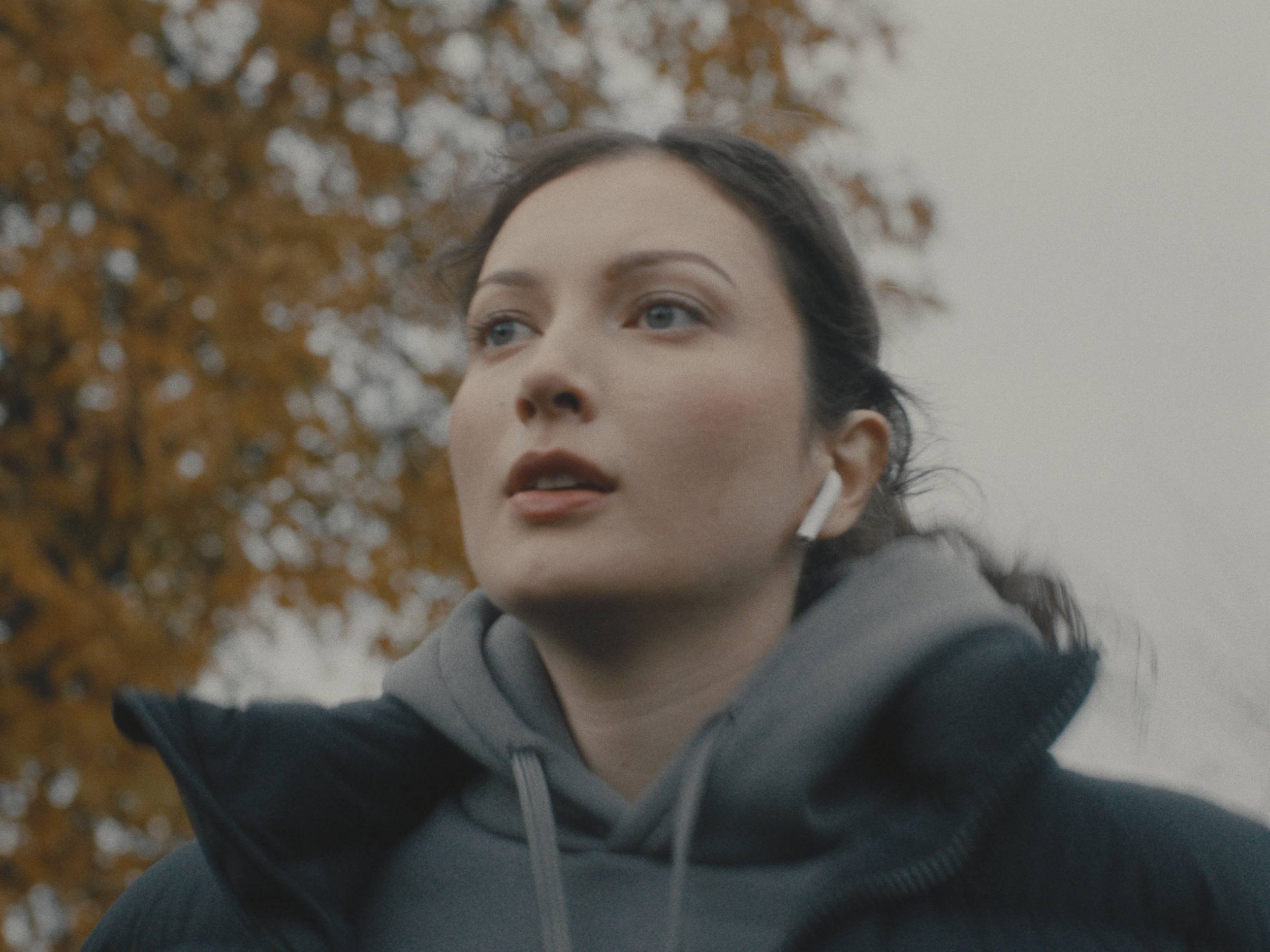 Close-up of a woman outdoors wearing a hoodie and jacket, with fall foliage in the background.
