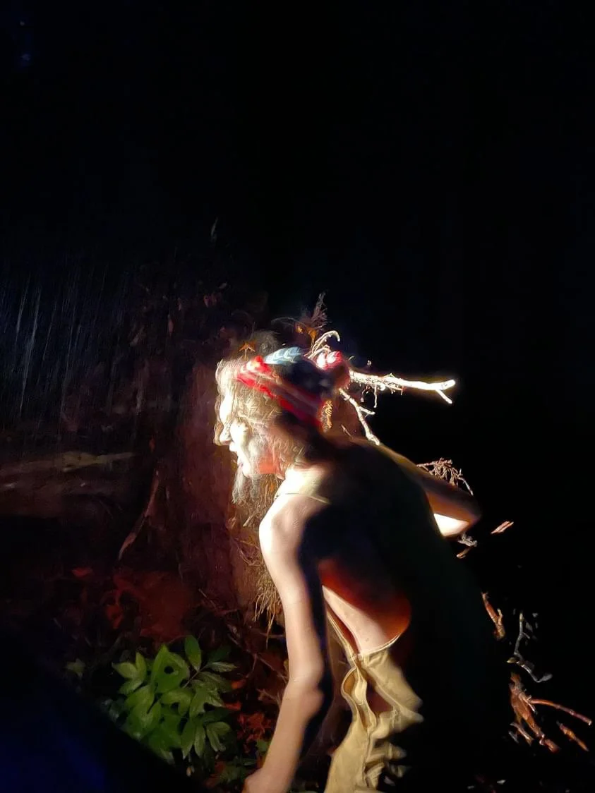 A person with long hair and a beard wearing a headdress with feathers, illuminated against a dark background, possibly at night or in a dimly lit environment.