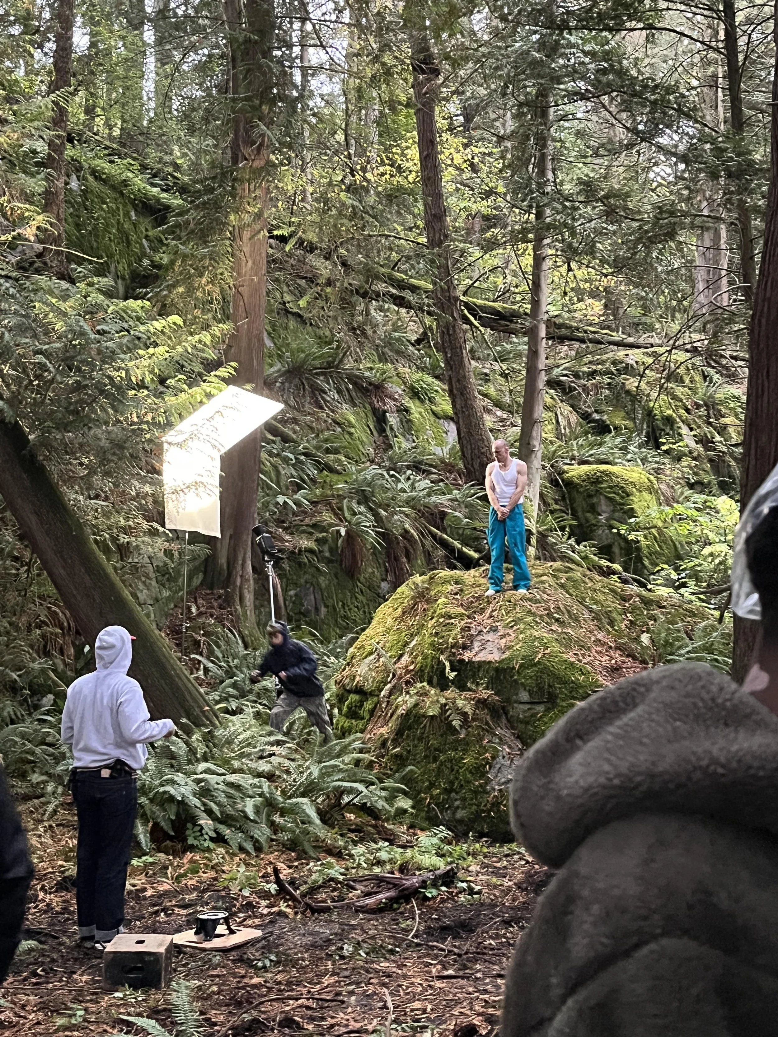 A photoshoot scene in a lush forest with trees and moss-covered rocks, featuring a man in blue pants standing on a large mossy rock, surrounded by crew members, some wearing hoodies, and filming equipment.