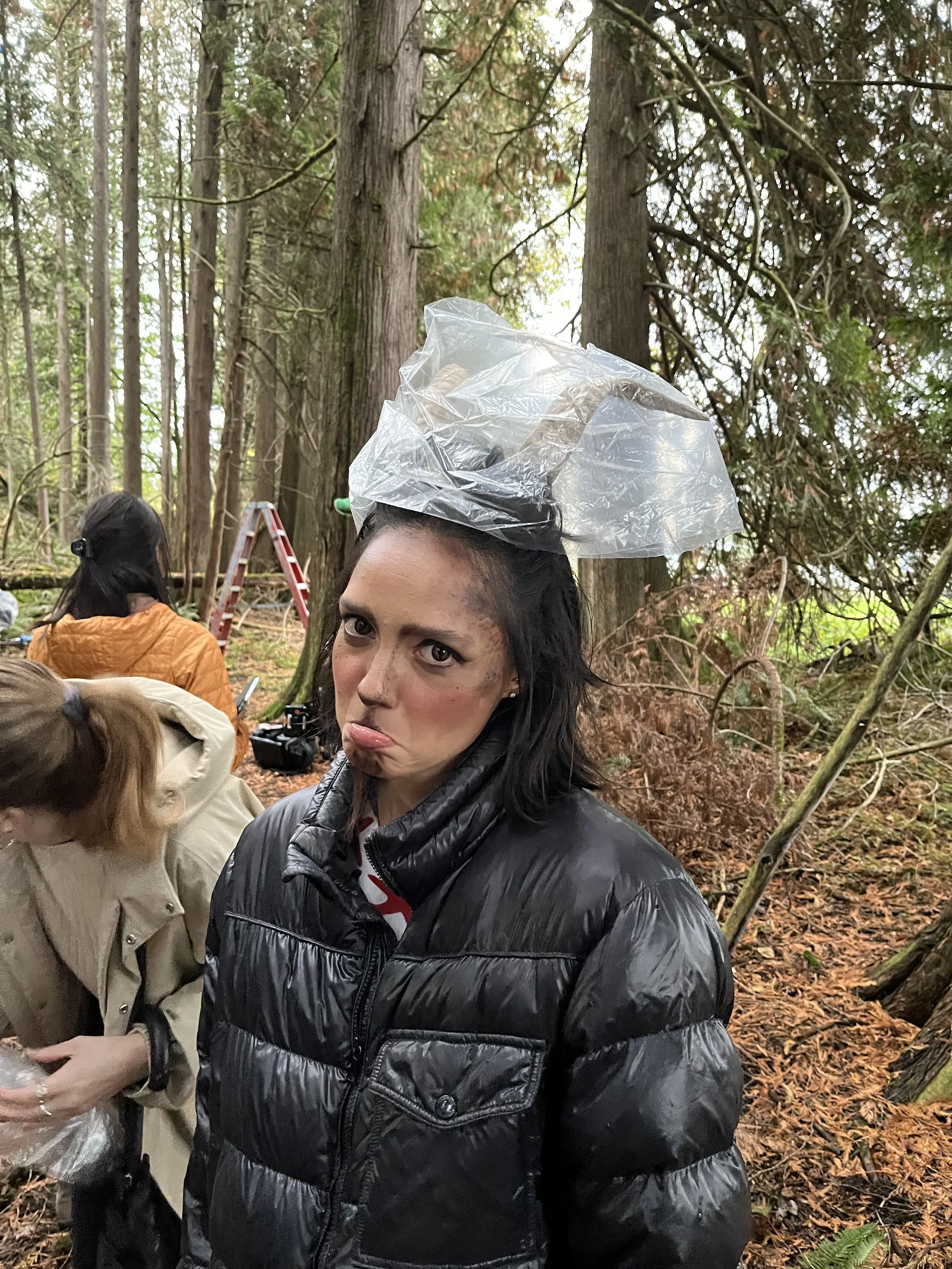 Woman with short black hair and a puffy black jacket in a forest, balancing a plastic bag on her head making a silly face, with other people and trees visible in the background.