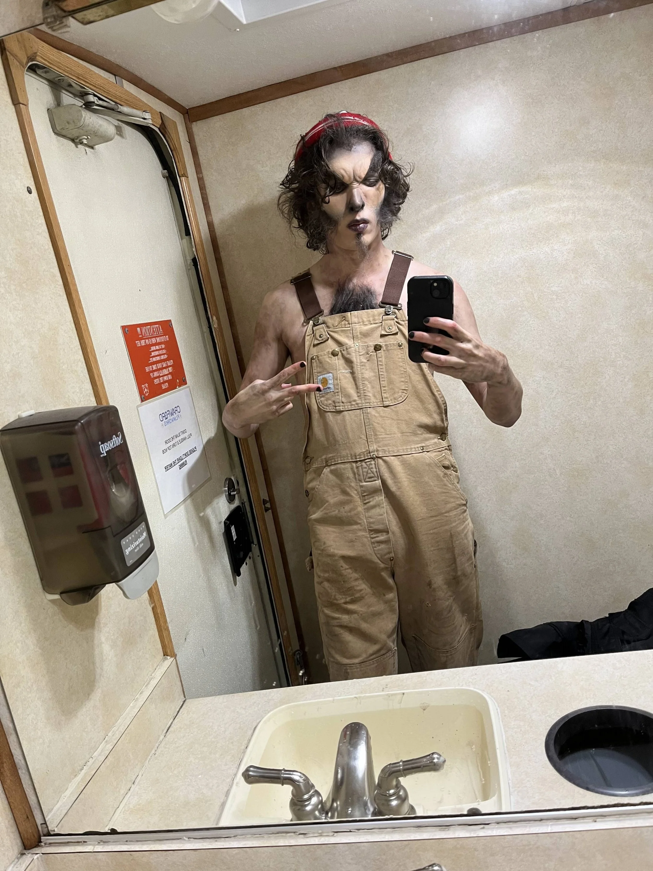 Person with makeup and curly hair taking a mirror selfie in a bathroom, wearing overalls with a red headband, pointing to a small clip on their overalls.
