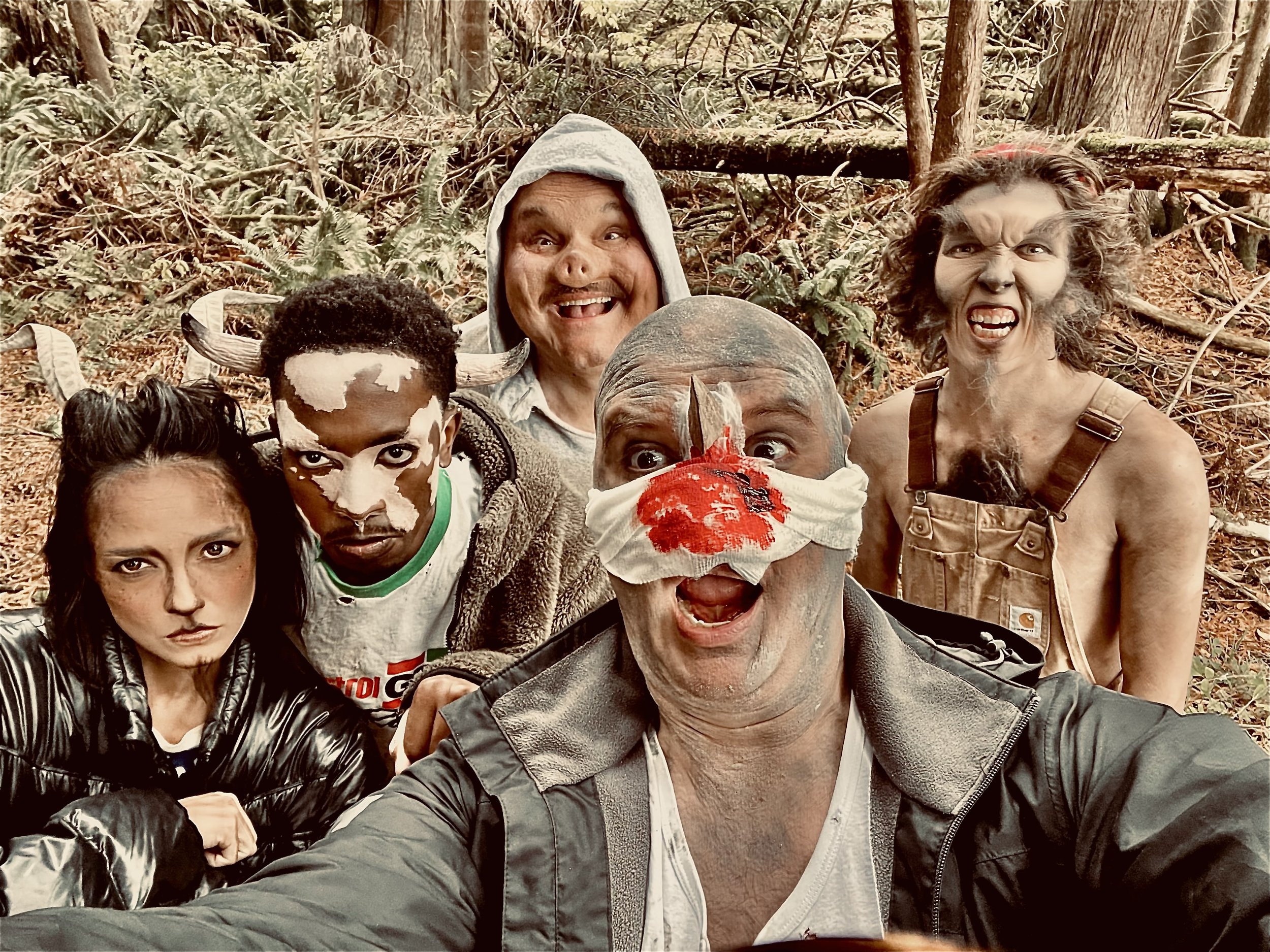 Group of people dressed in horror or fantasy costumes, including a person with a bloody bandaged face and horns, a person with painted face and cow print, a person with a bloodstained nose, another with zombie makeup, and a person with wild hair and 