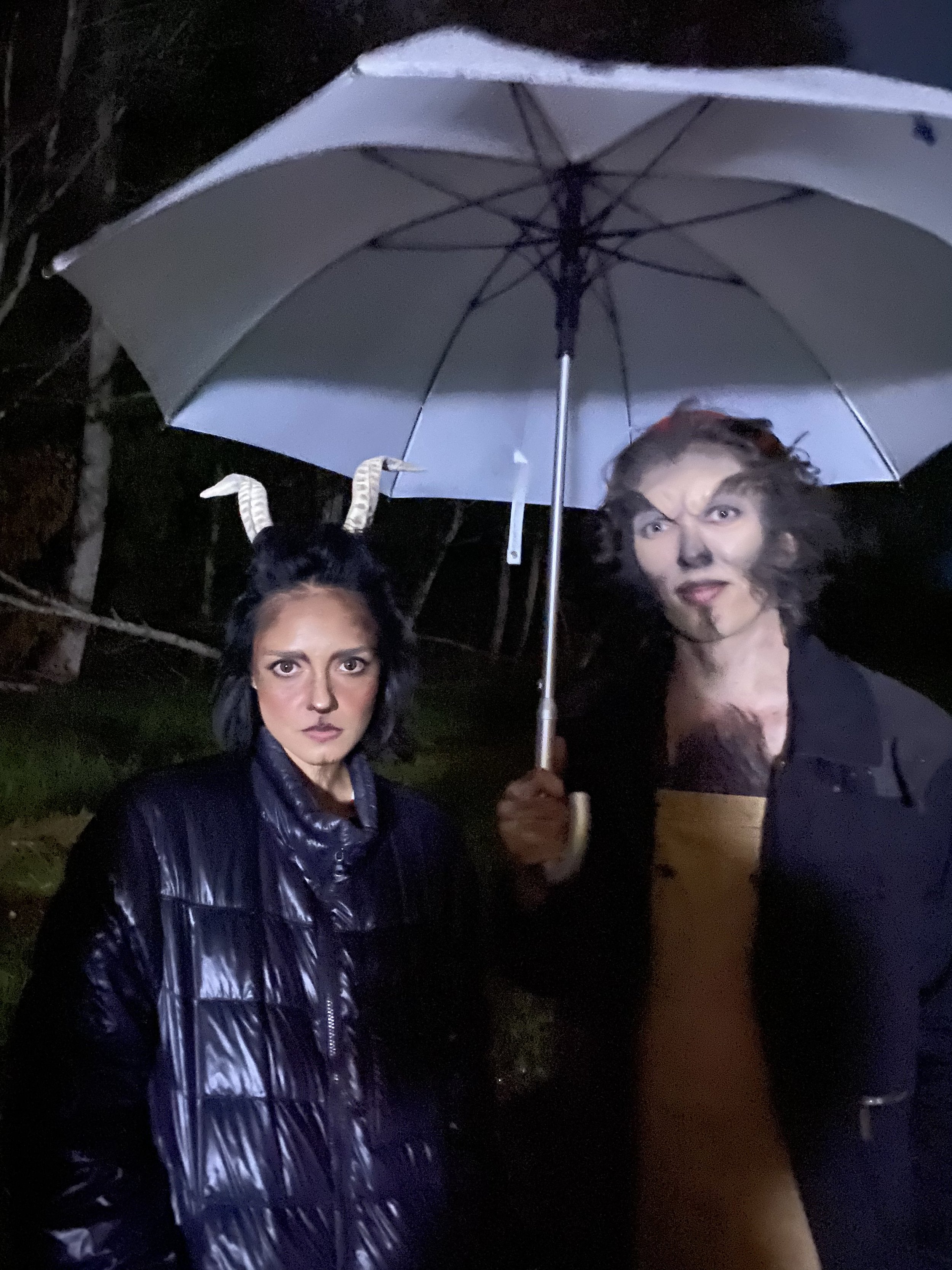 Two people standing outdoors at night under a large umbrella, with one person wearing horn-like headwear and the other making a face with wild hair.