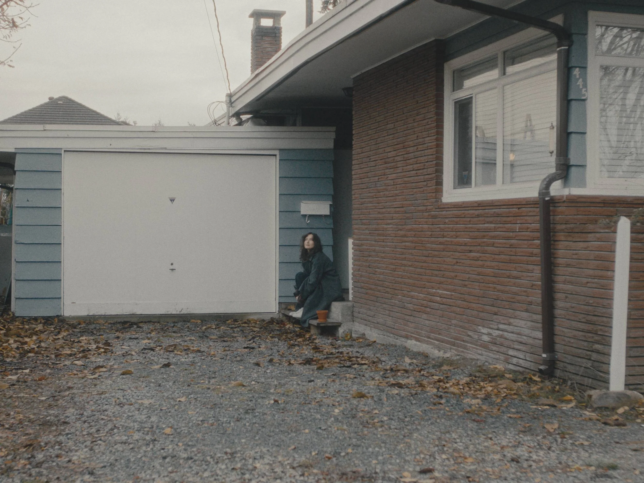 A woman with long dark hair wearing a dark coat is sitting on the step of a house, next to a small empty flower pot, in front of a blue garage door. The house has a brick facade and a window with horizontal blinds. There are fallen leaves on the grav