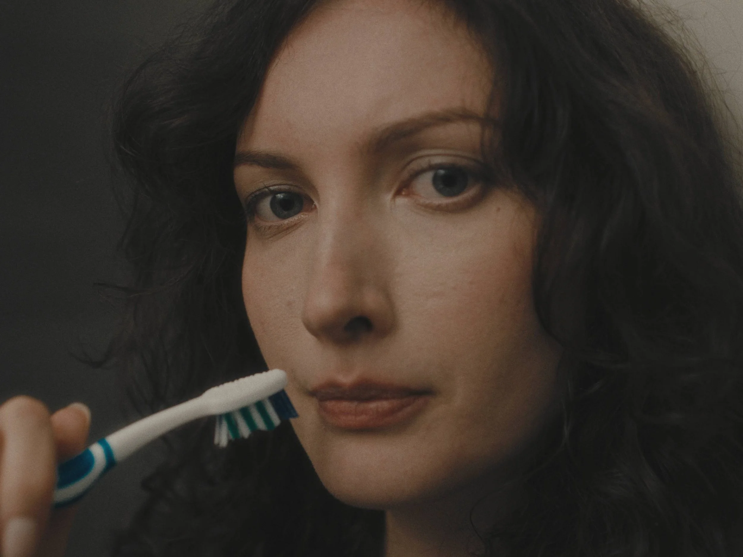 A woman with black, curly hair looking at the camera while holding a toothbrush near her lips.