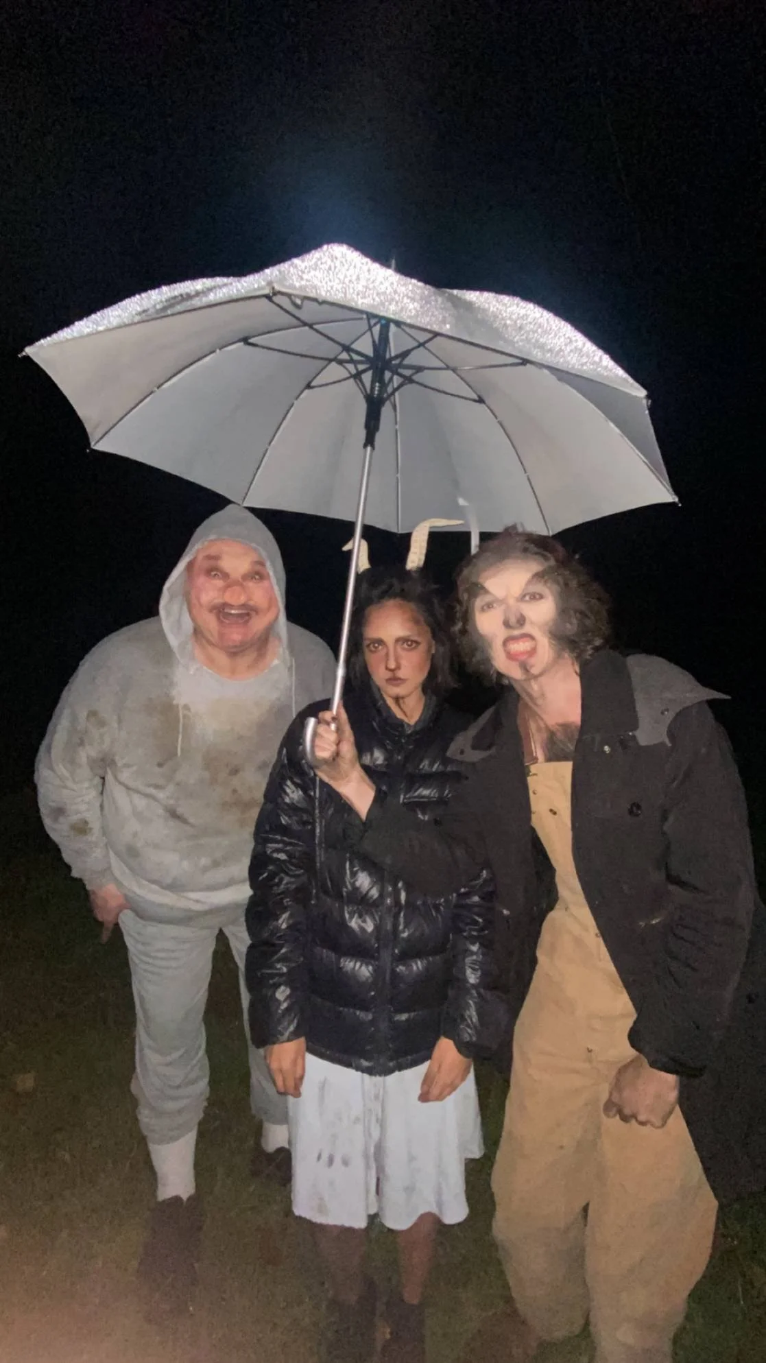Three people dressed in costumes standing outside at night under an umbrella, making funny faces.