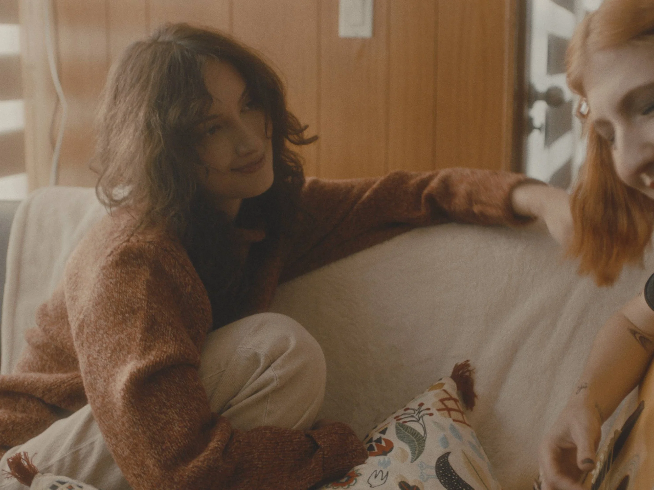 A woman with wavy brown hair, wearing a brown sweater, sitting on a cream sofa in a room with wooden walls, leaning her arm on the back of the sofa, smiling softly.