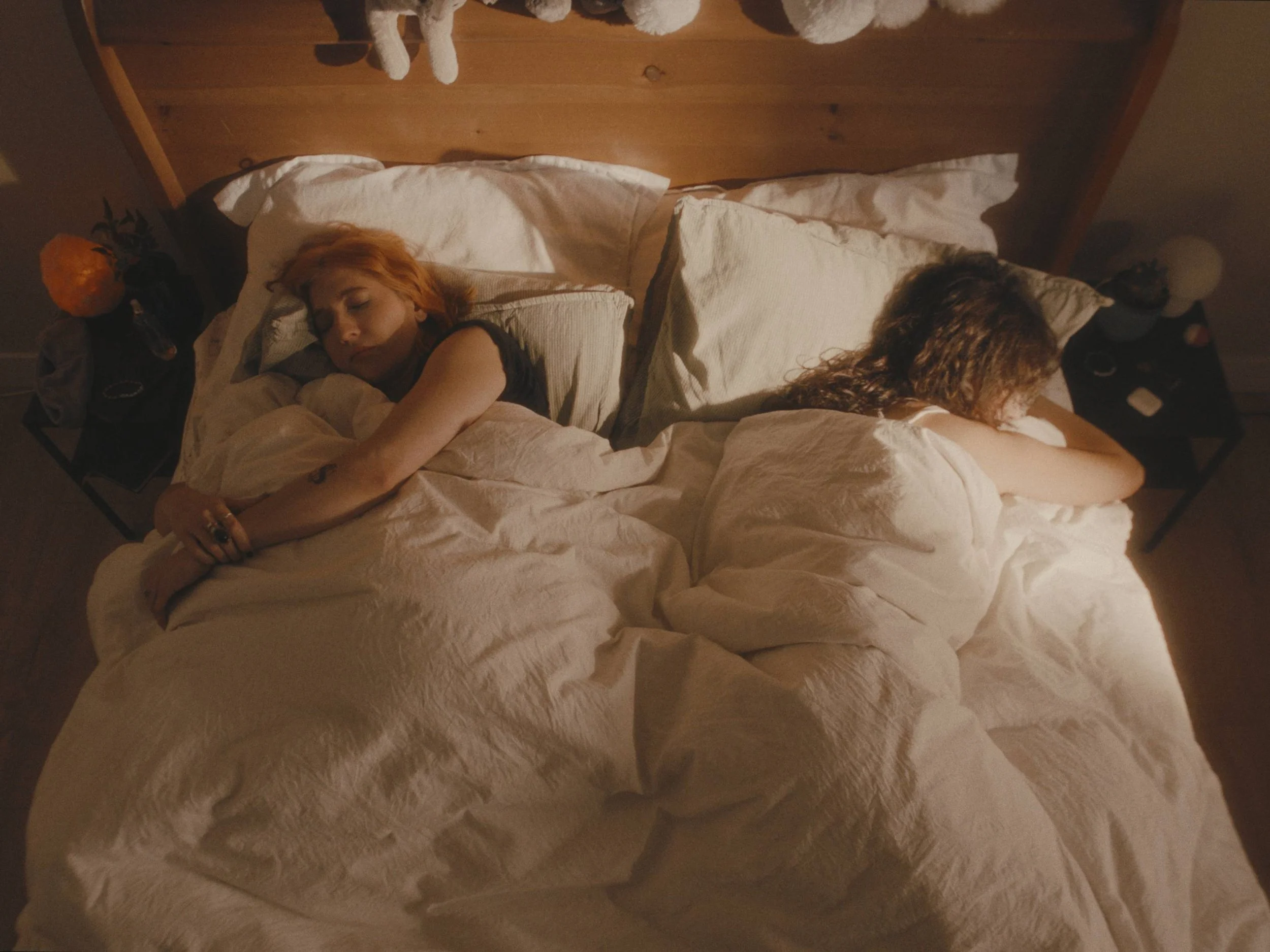 Two women are sleeping in bed, lying on opposite sides with their backs to each other, with a wooden headboard and side tables with lamps and personal items.