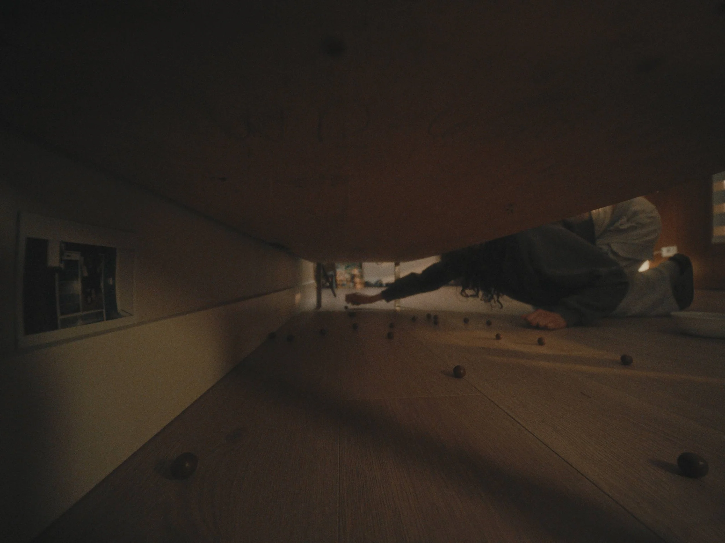 Person lying on the floor reaching towards objects scattered on the ground, viewed from underneath a table or furniture.