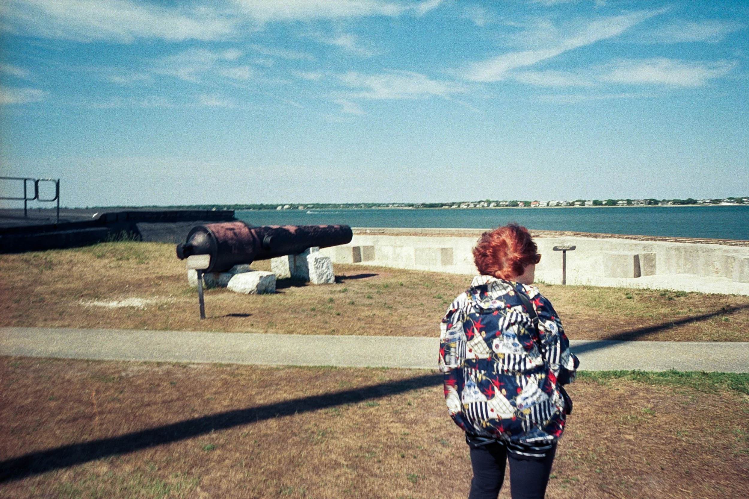 A woman wearing a colorful jacket at Fort Sumter with a canal or river in the background. There is a rusted cannon on a concrete platform and benches nearby. The sky is mostly clear with some clouds.