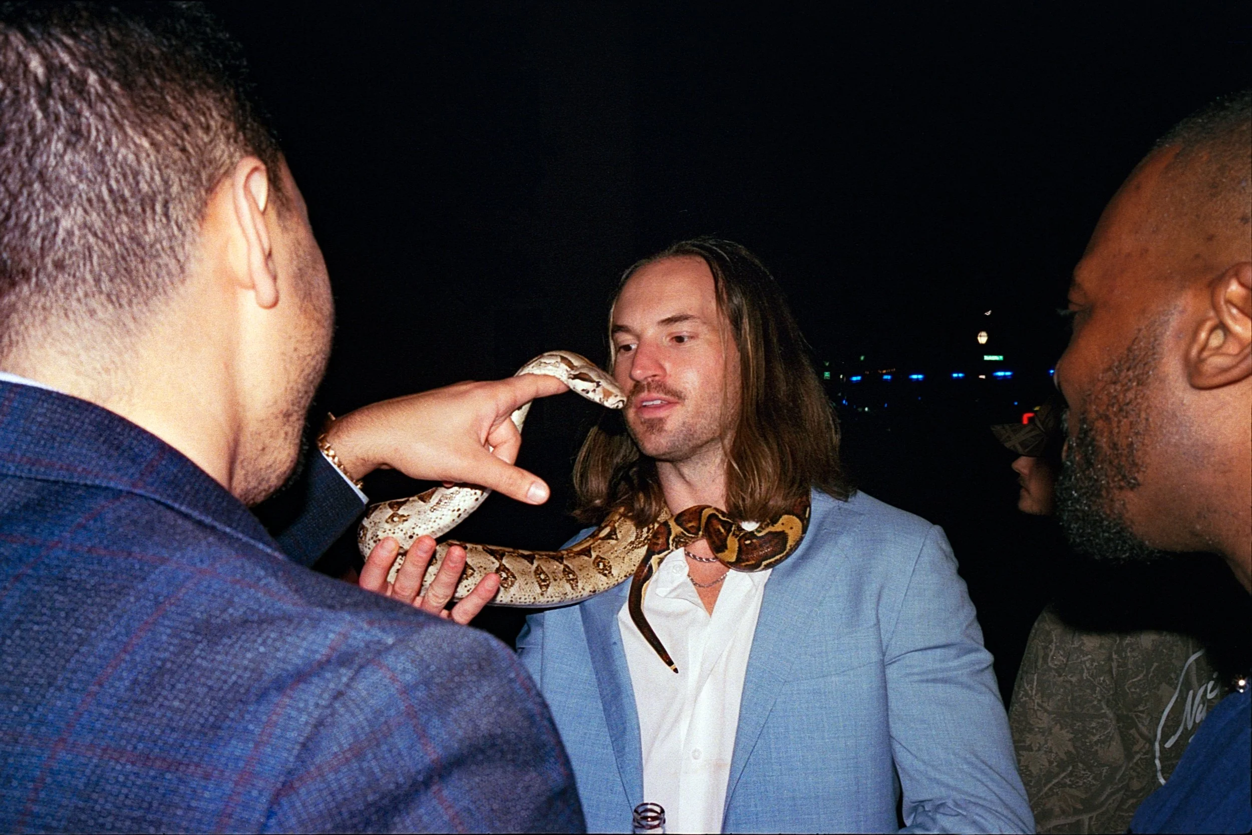 Three men are at a party, one holding a snake close to another man's face, who is looking at it with a slight smile.