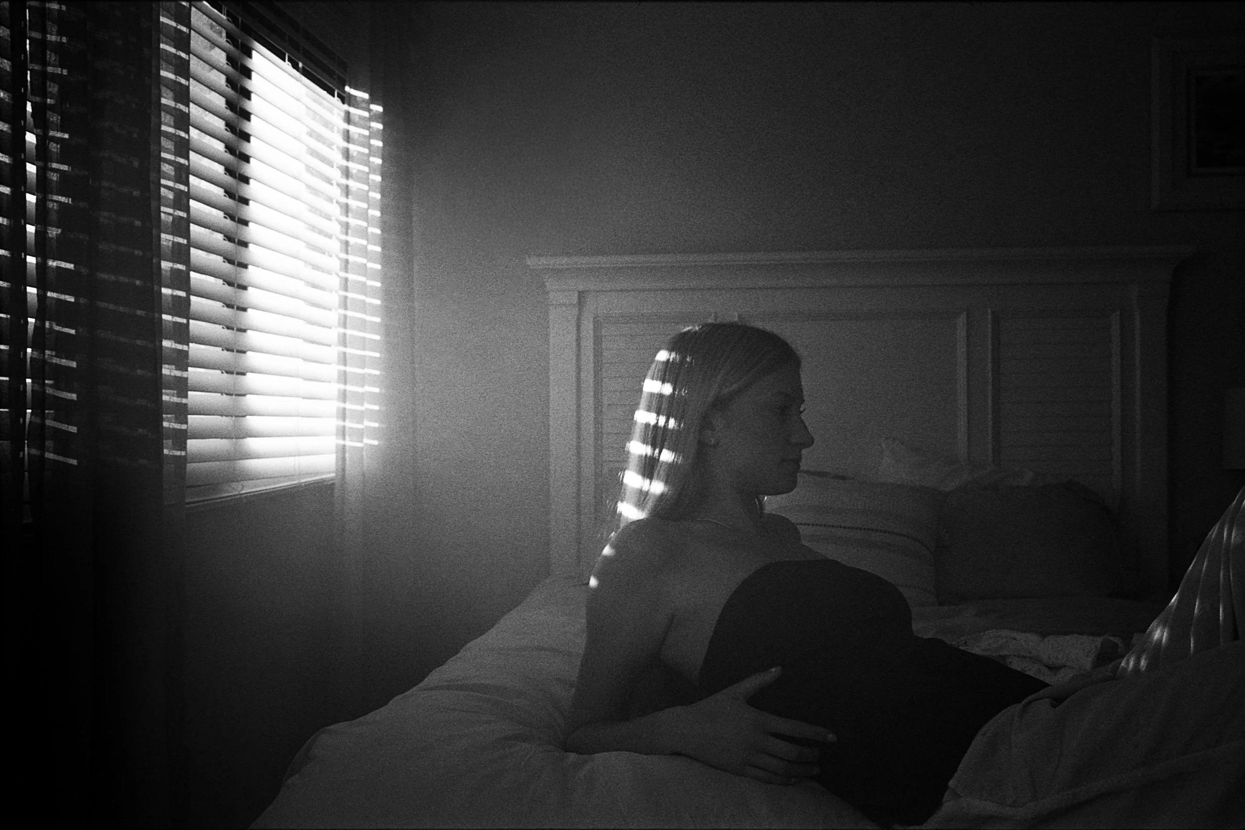 A black and white photo of a woman with long hair sitting in a room with sunlight coming through closed blinds, casting striped shadows on her and the wall.