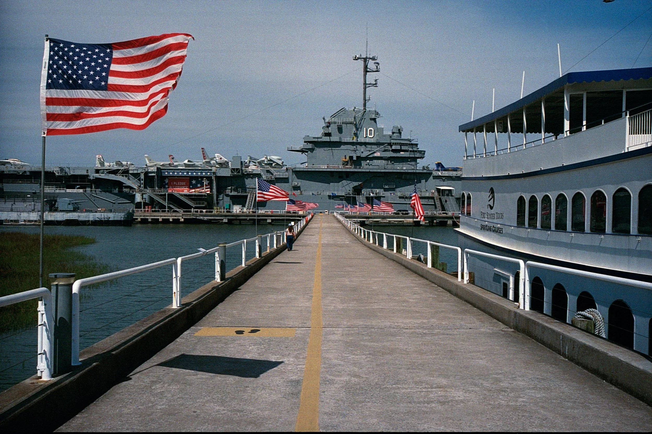 A marina with American flags, The USS YORKTOWN is in the distance , smaller planes on the ship's deck, and a boat on the water labeled 'Two Sister Tours - Sightseeing Cruises' docked at the pier.