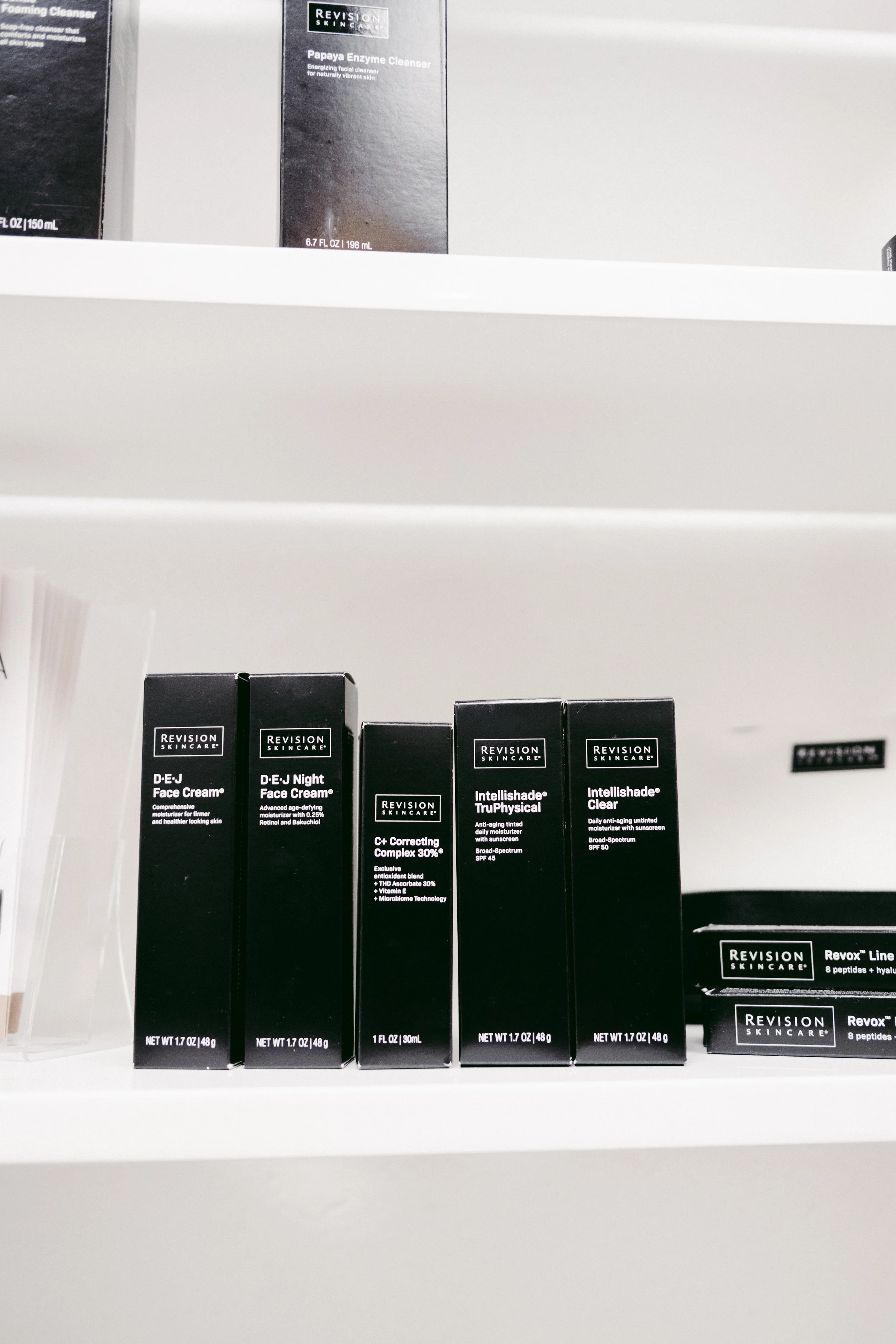 Shelf displaying various skincare products, including Revision Skincare items such as D.E.j Face Cream, D.E. Night Face Cream, C+ Correcting Complex 30%, Intellishade TruPhysical, and Intellishade Clear, with mostly black packaging and white text.