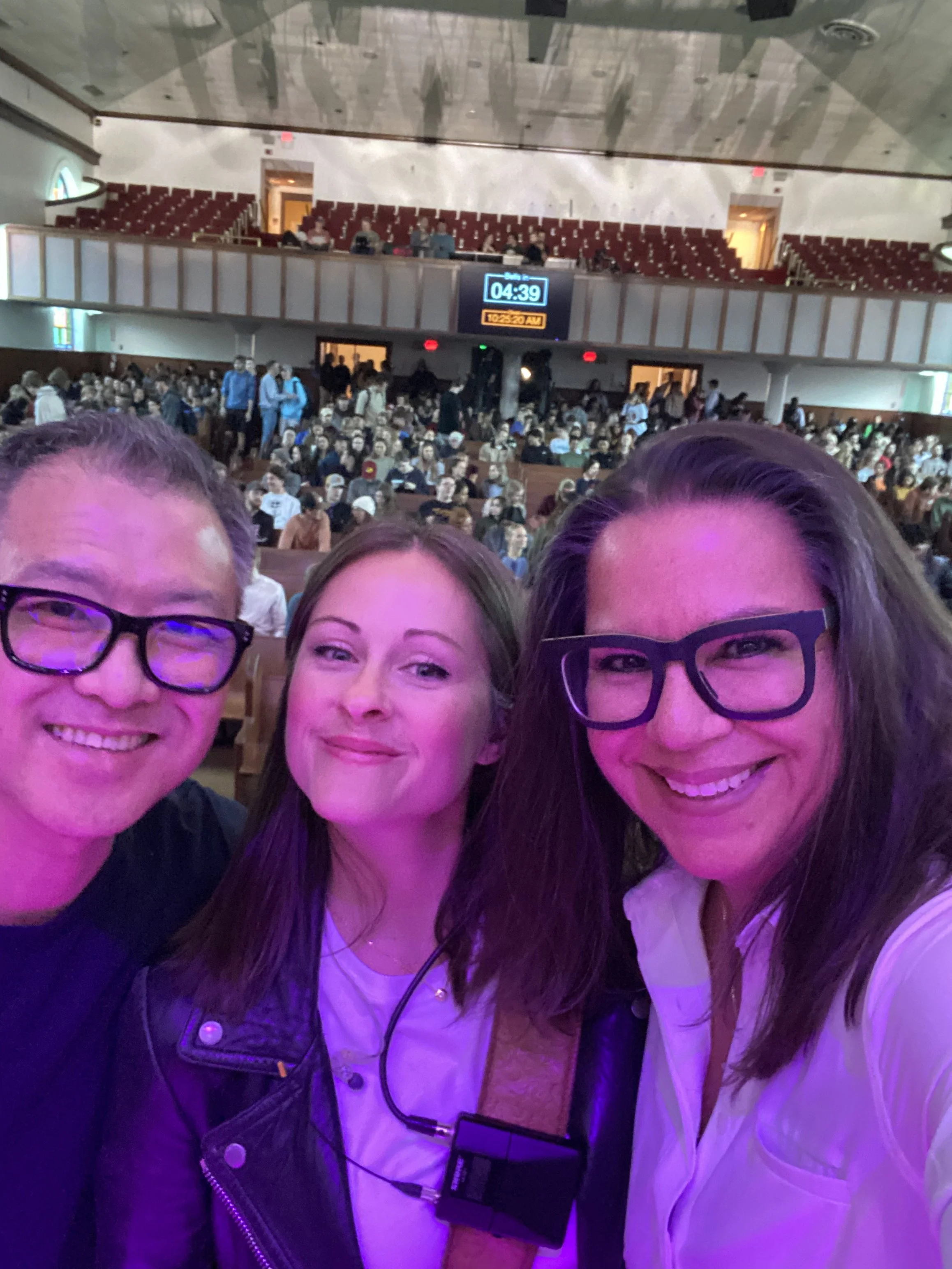 Three people in the foreground taking a selfie inside a large auditorium filled with people seated and standing