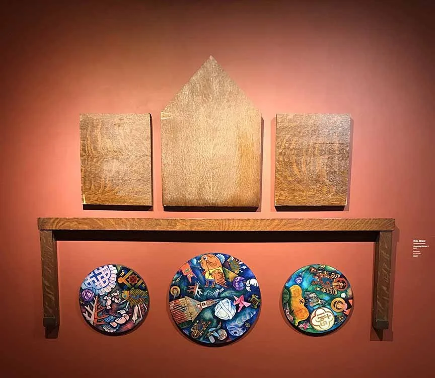 An art display with three wooden panels at the top and a wooden shelf beneath them, holding three colorful circular paintings on a red wall.
