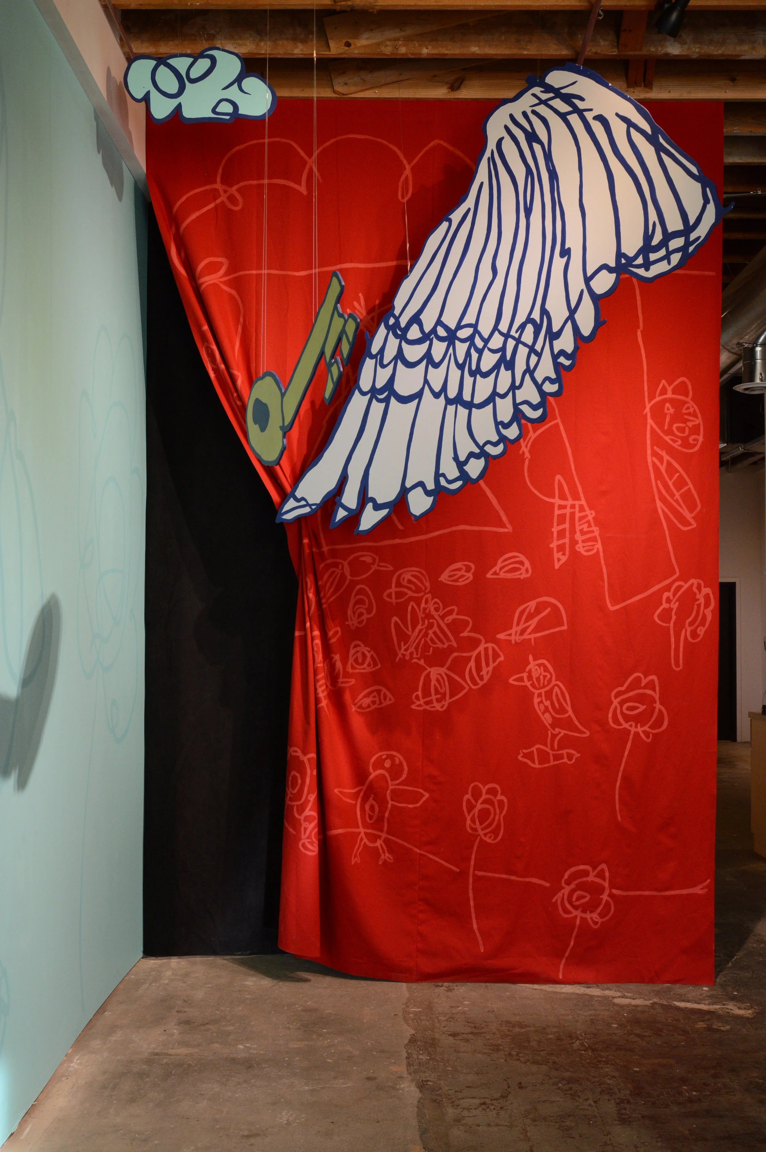 A colorful art display with a large, stylized illustration of a wing, a key, and various sketch-like drawings of animals and flowers on a red curtain backdrop, with a light blue wall to the side.