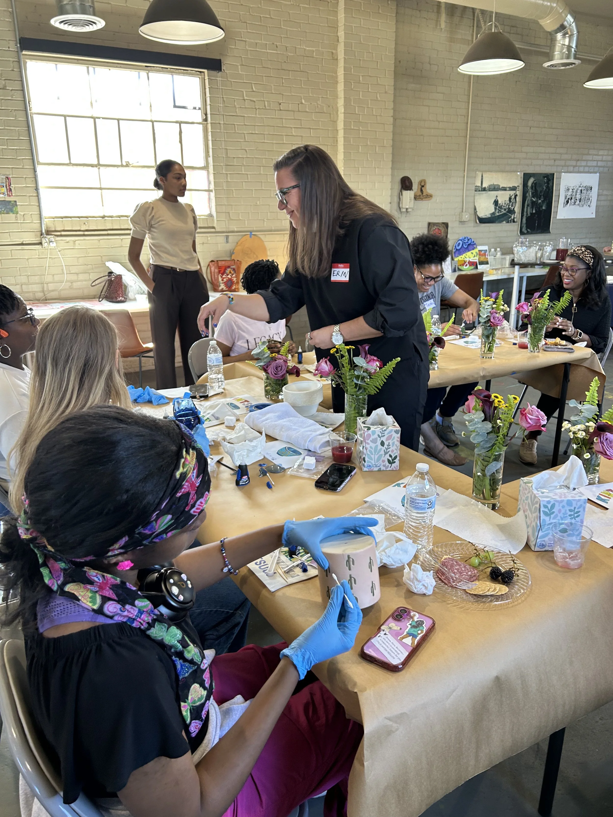 Group of women participating in a workshop session in a well-lit room with brick walls. The women are seated at a long table with floral arrangements and craft supplies, while one woman stands and demonstrates.