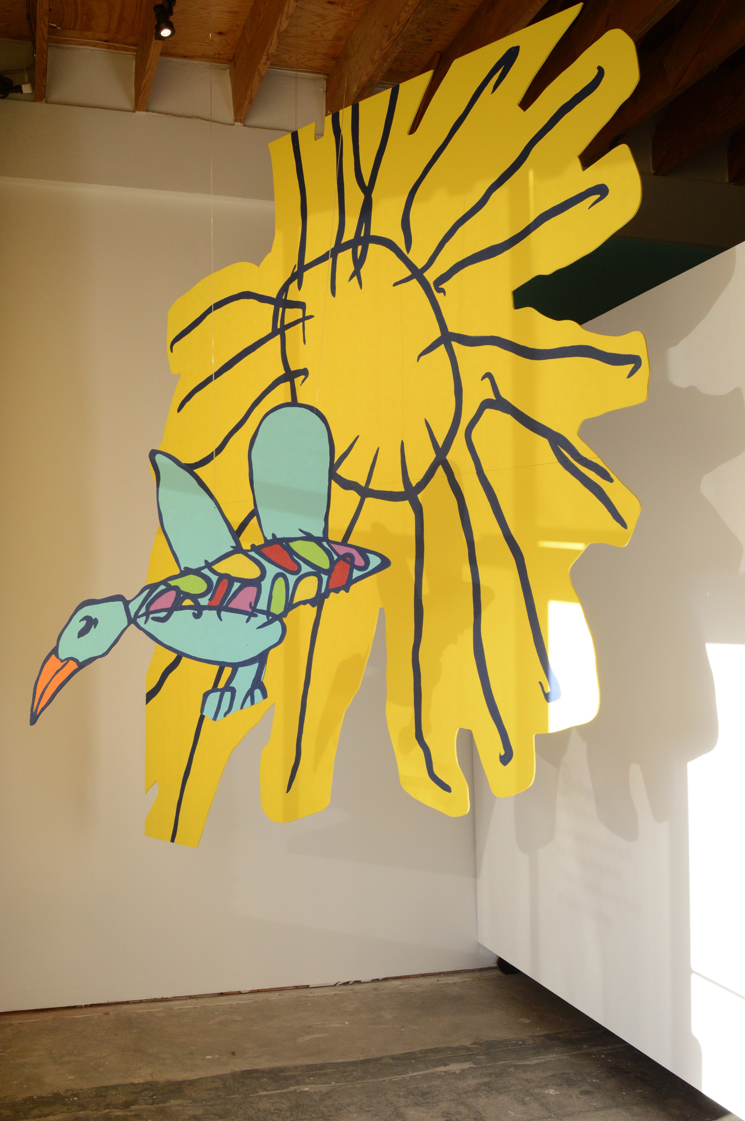 A large yellow cutout of a sunflower with black outline details, painted on the wall with a stylized bird drawn in bright pastel colors flying near it.