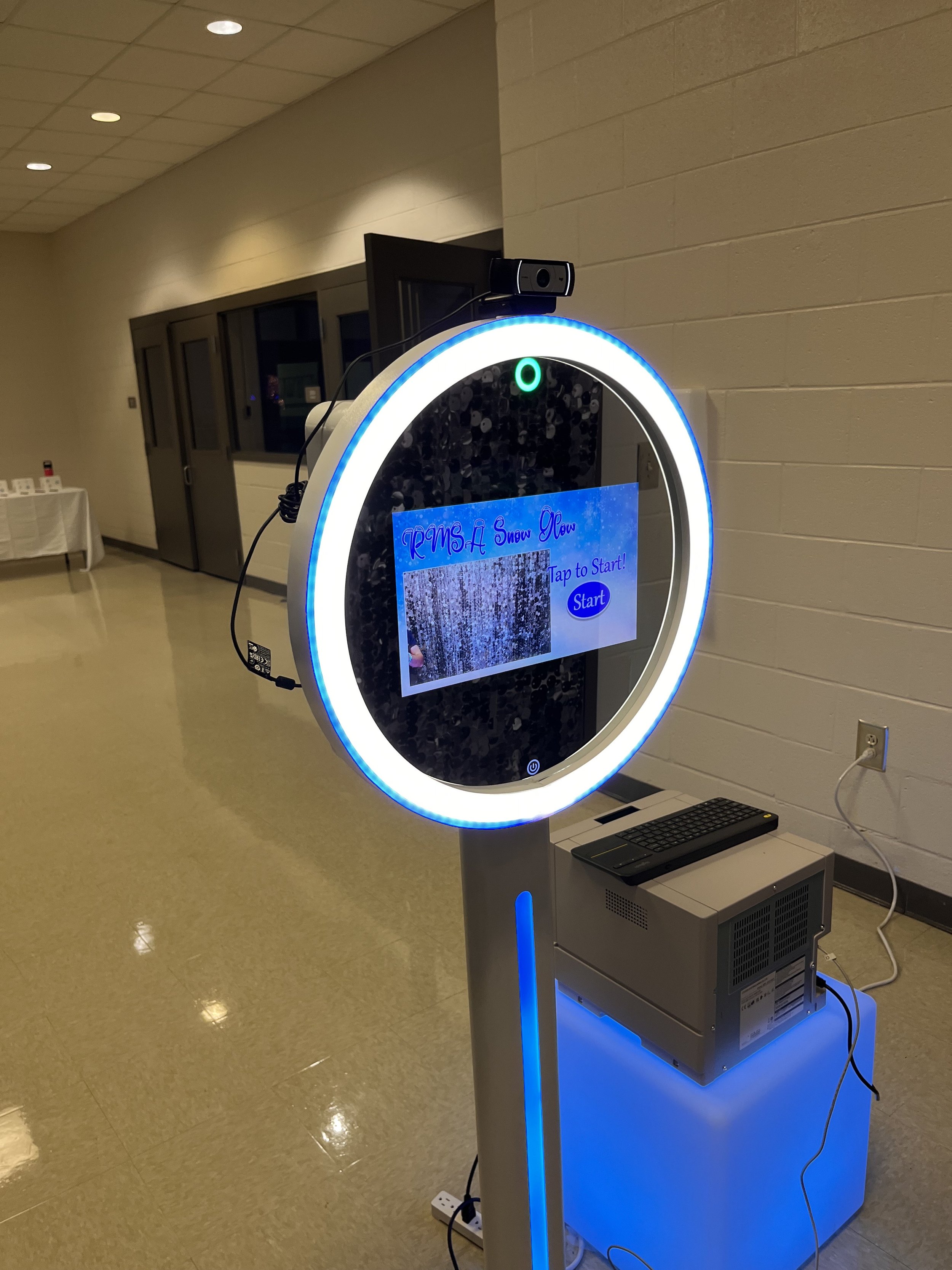 A circular touchscreen photo booth with a blue LED light ring, displaying an invite to start a snow-themed photo session, situated in an indoor hallway.