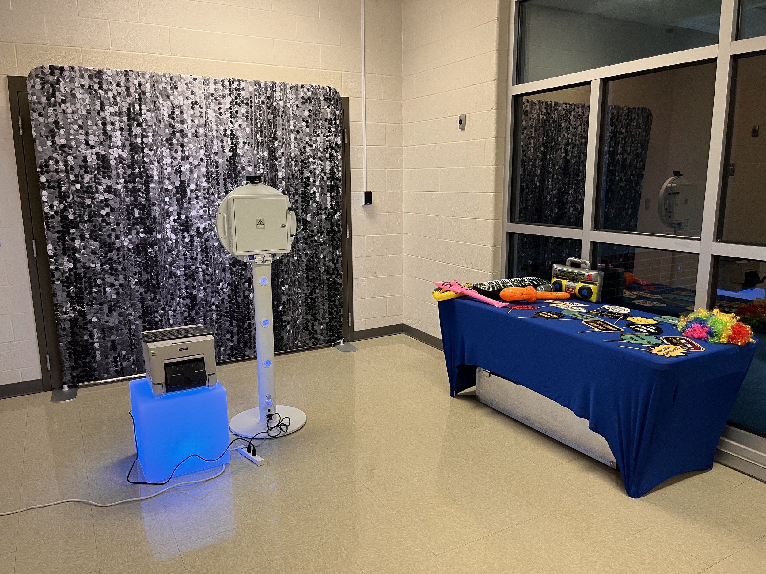 Photo of a photo booth setup with a black sparkle backdrop, a white photo booth camera, and a table with party props including leis, sunglasses, and boombox cutouts.