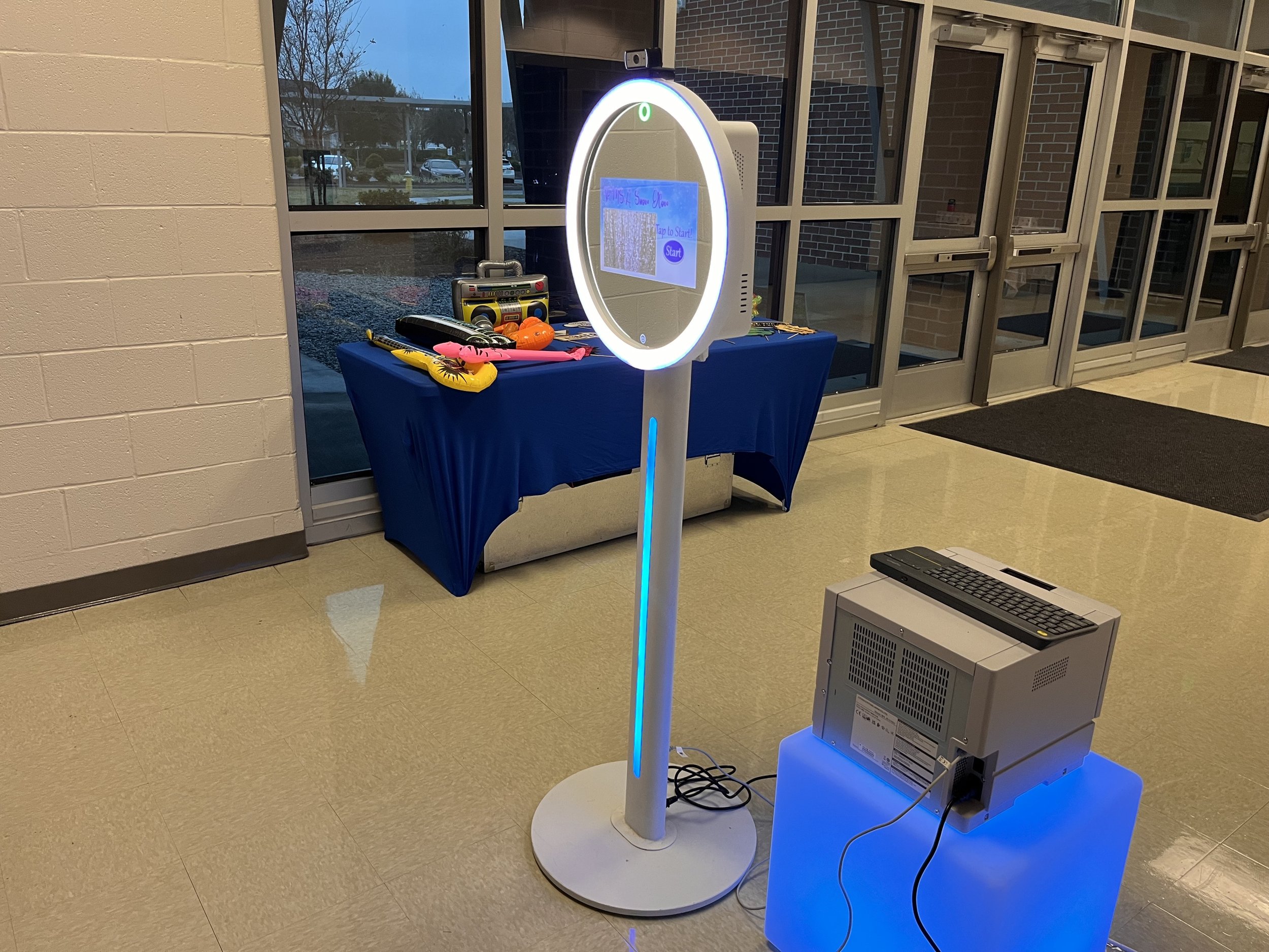 A selfie station setup in an indoor space with a ring light, touchscreen, and a printer or scanner on a blue-lit stand. In the background, there is a table with balloons and a music player.