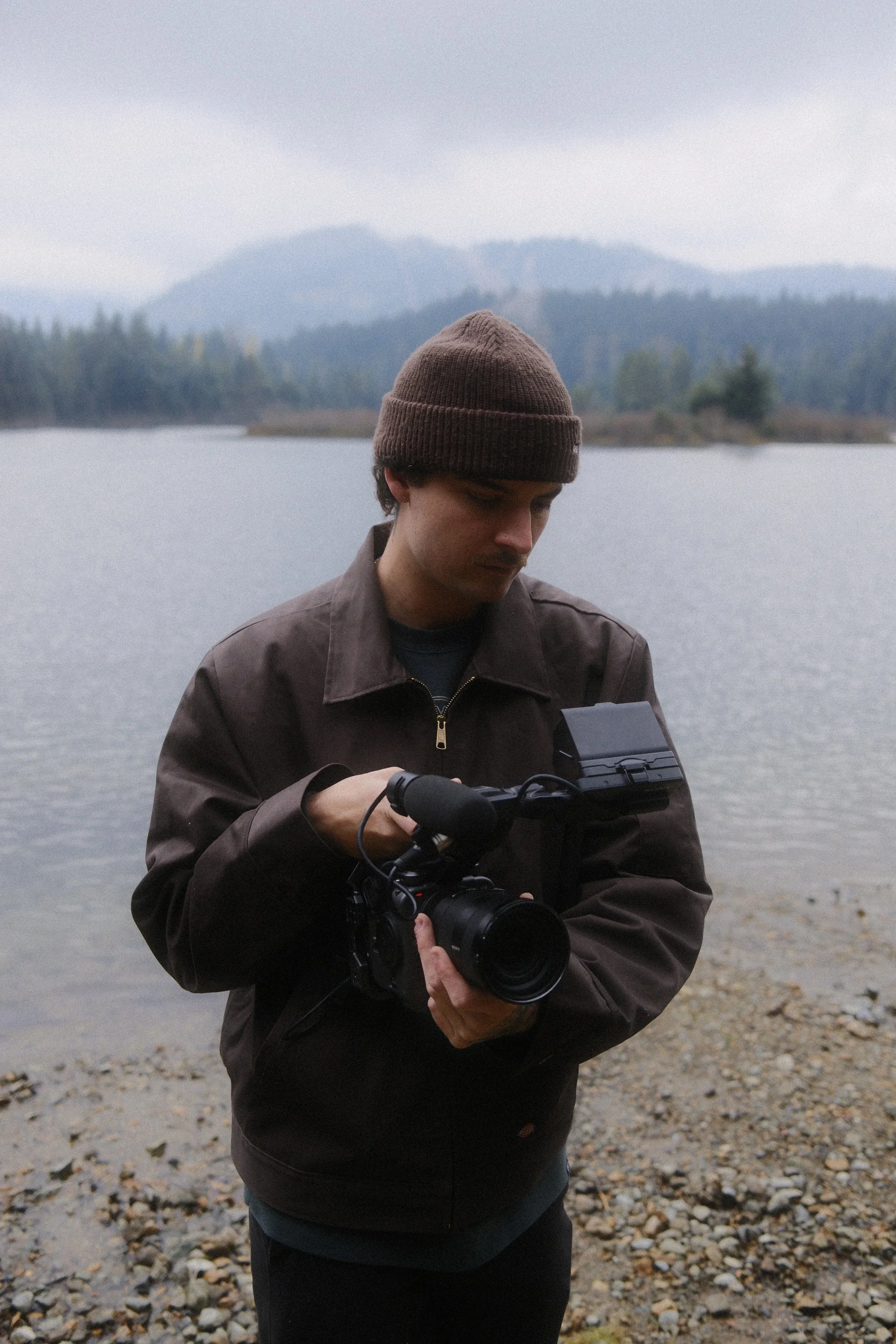 A man wearing a brown knit beanie and dark jacket standing by a lake, holding a camera in his hands, with mountains and trees in the background.