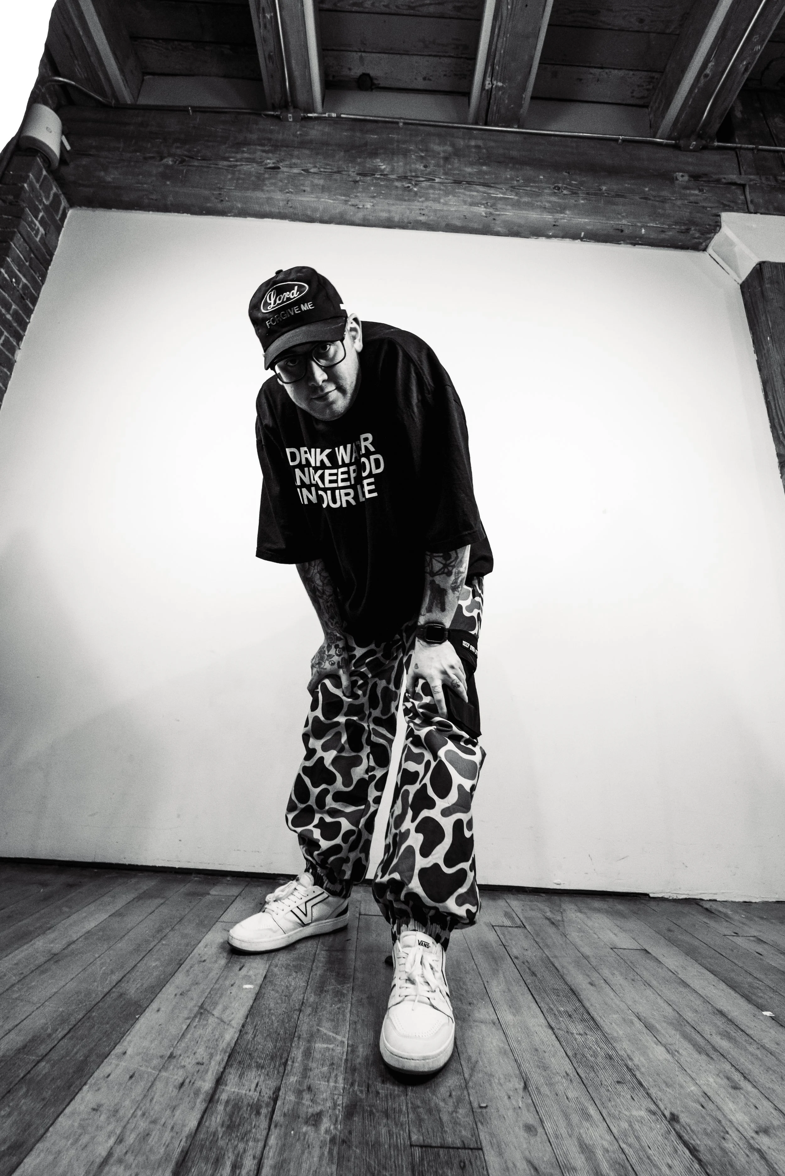 A young man standing indoors against a plain white wall, wearing glasses, a black cap, a black T-shirt with white text, camouflage cargo pants, and white sneakers.