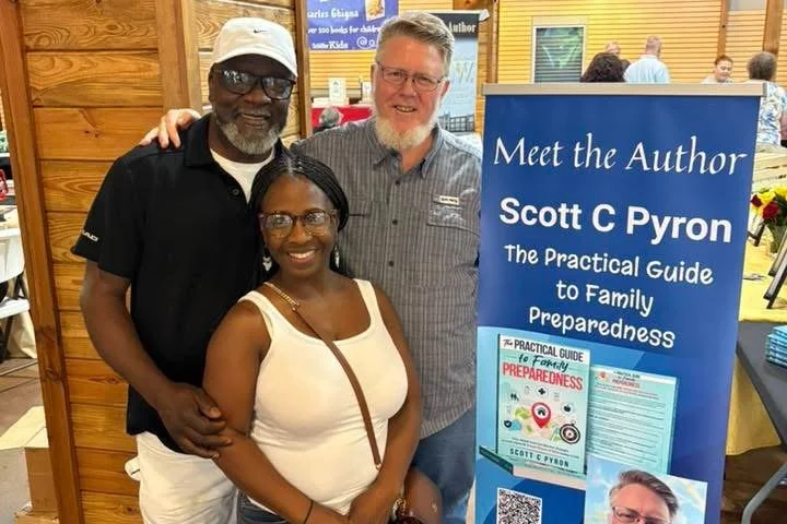 Three people, including Scott C Pyron, stand together for a photo at a book signing event. A blue sign features their names and book title, 'The Practical Guide to Family Preparedness.'