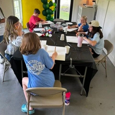 Children sitting around a table engaged in a craft activity with paper, markers, and cups in a room decorated with green balloons.