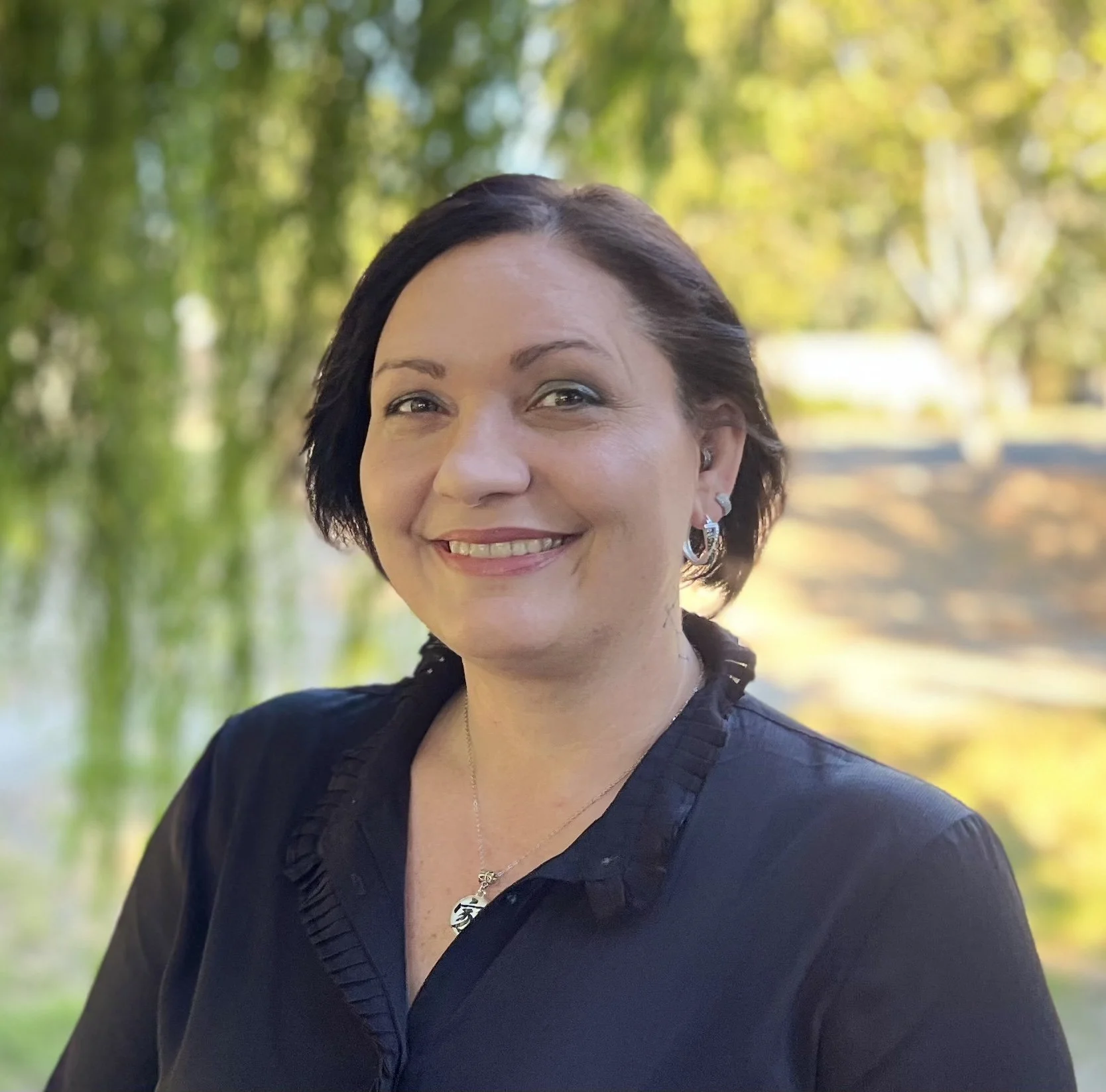 A smiling woman with short dark hair outdoors, wearing earrings and a necklace, with a blurred background of green and yellow trees.