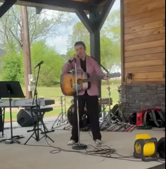 A man playing an acoustic guitar and singing into a microphone on an outdoor stage with musical equipment around him, including a keyboard, drum set, and speakers. The stage is under a wooden pavilion with trees visible in the background.