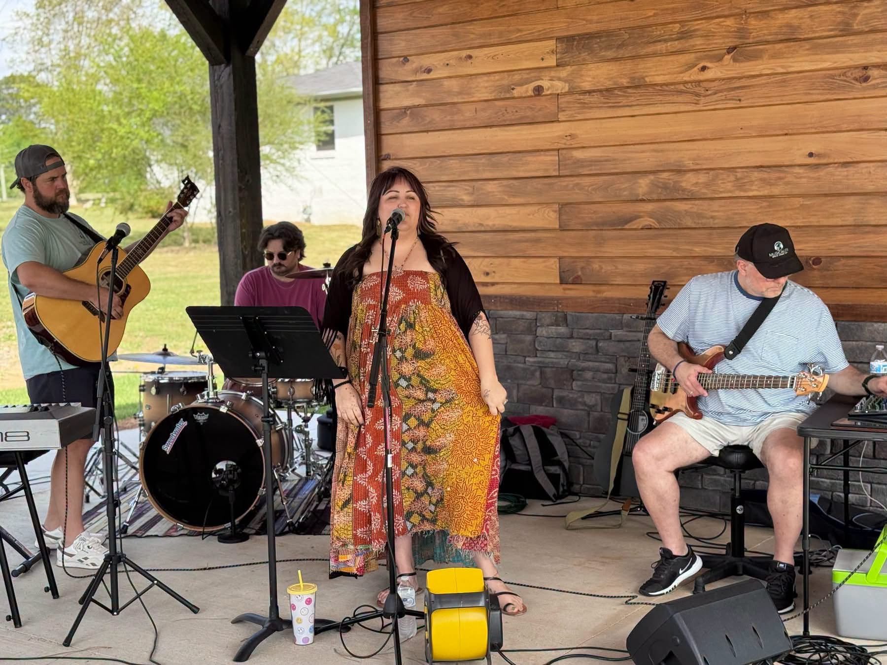 A band performing outdoors under a pavilion, with four musicians including a woman singer, a guitarist, a drummer, and another guitarist, against a backdrop of green trees and a white house.