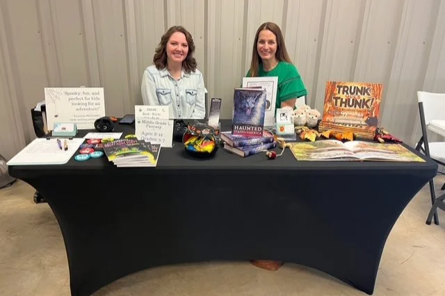 Two women smiling behind a table with books, pamphlets, and Halloween decorations at an indoor event.