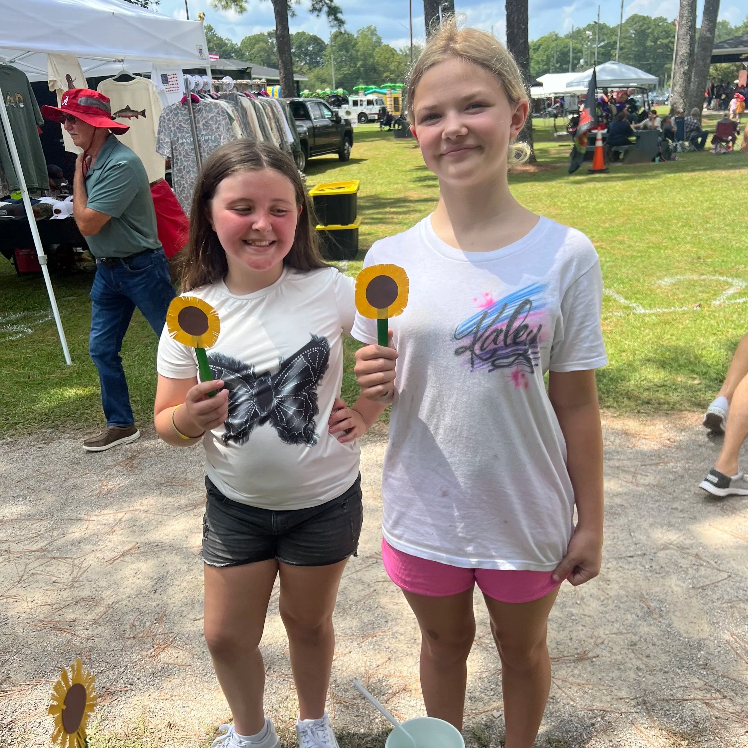 Two young girls standing outdoors at a fair or festival, holding yellow and brown sunflower-shaped objects, with booths and tents in the background containing clothing and other items, and people sitting and walking around.