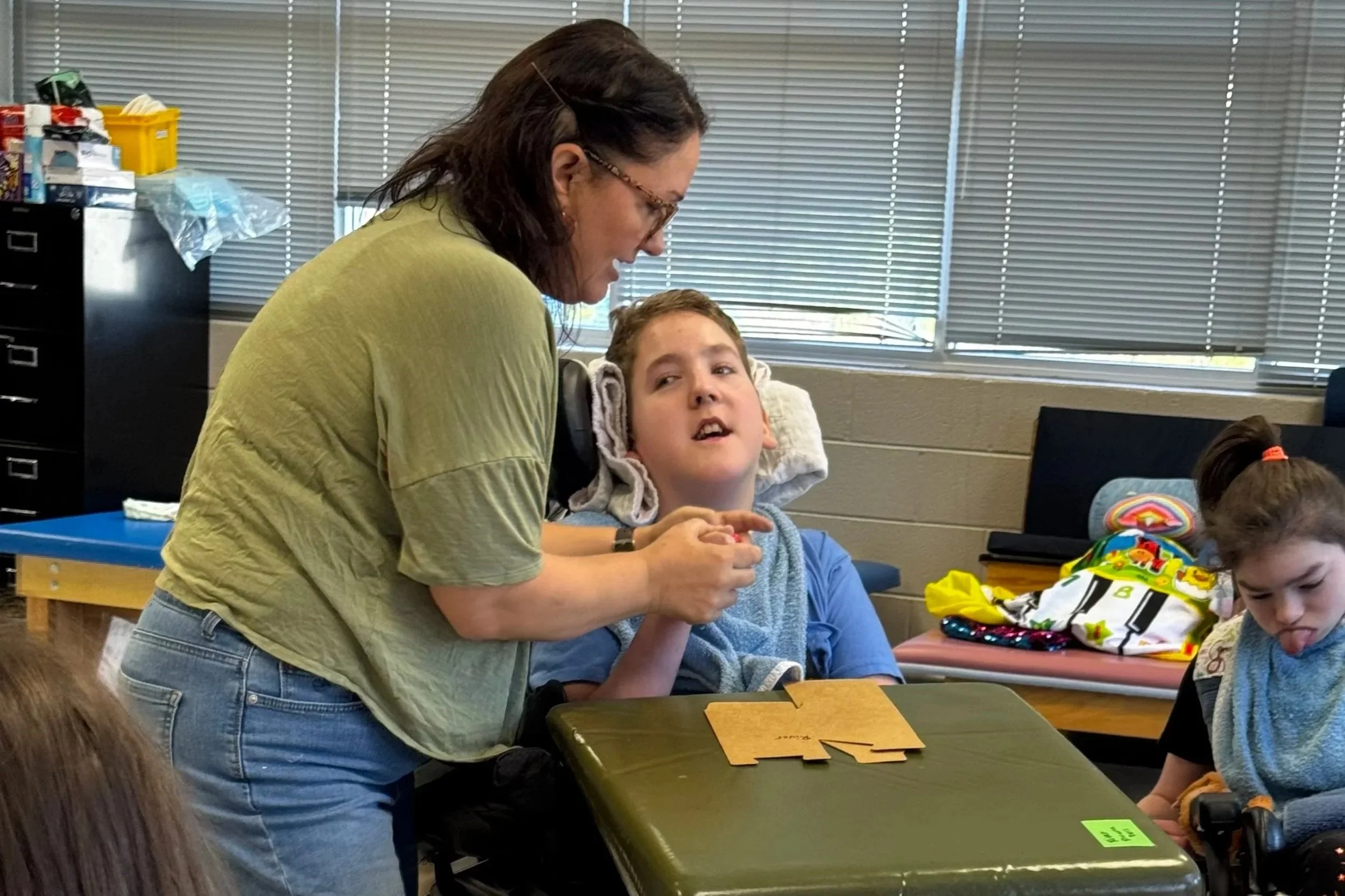 A woman with dark hair and glasses leaning over a young boy in a wheelchair, playing an educational game at a classroom table. Several children are sitting at the table, with school supplies and colorful bags visible, in a room with large windows and closed blinds.