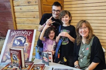 A group of four people at a table with Werewolf Hamlet graphic novels and merchandise, smiling and making peace signs.