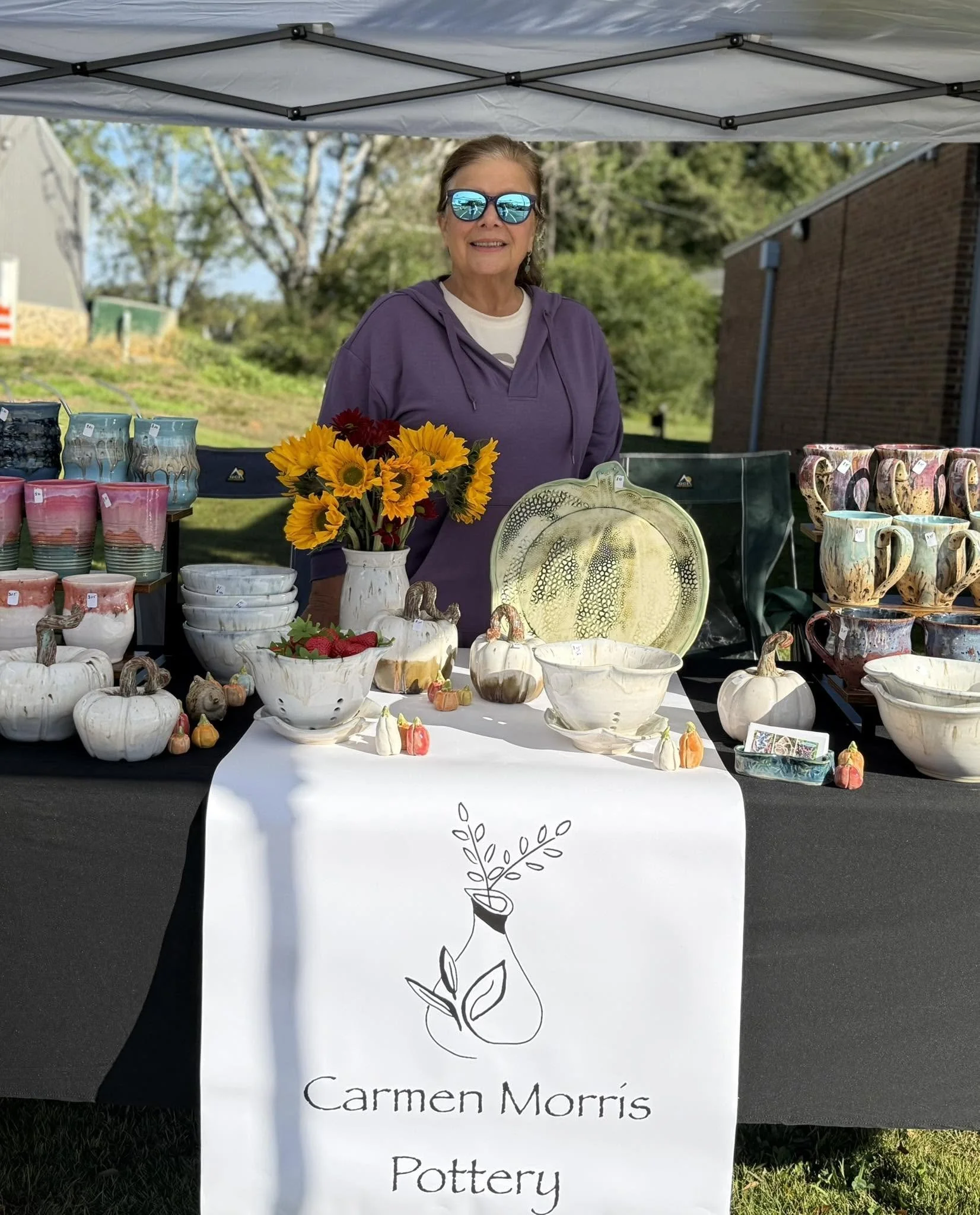 The image shows a woman standing behind a table at an outdoor pottery stand under a canopy, with various bowls, mugs, and pumpkins on display, and a sign that reads "Carmen Morris Pottery."