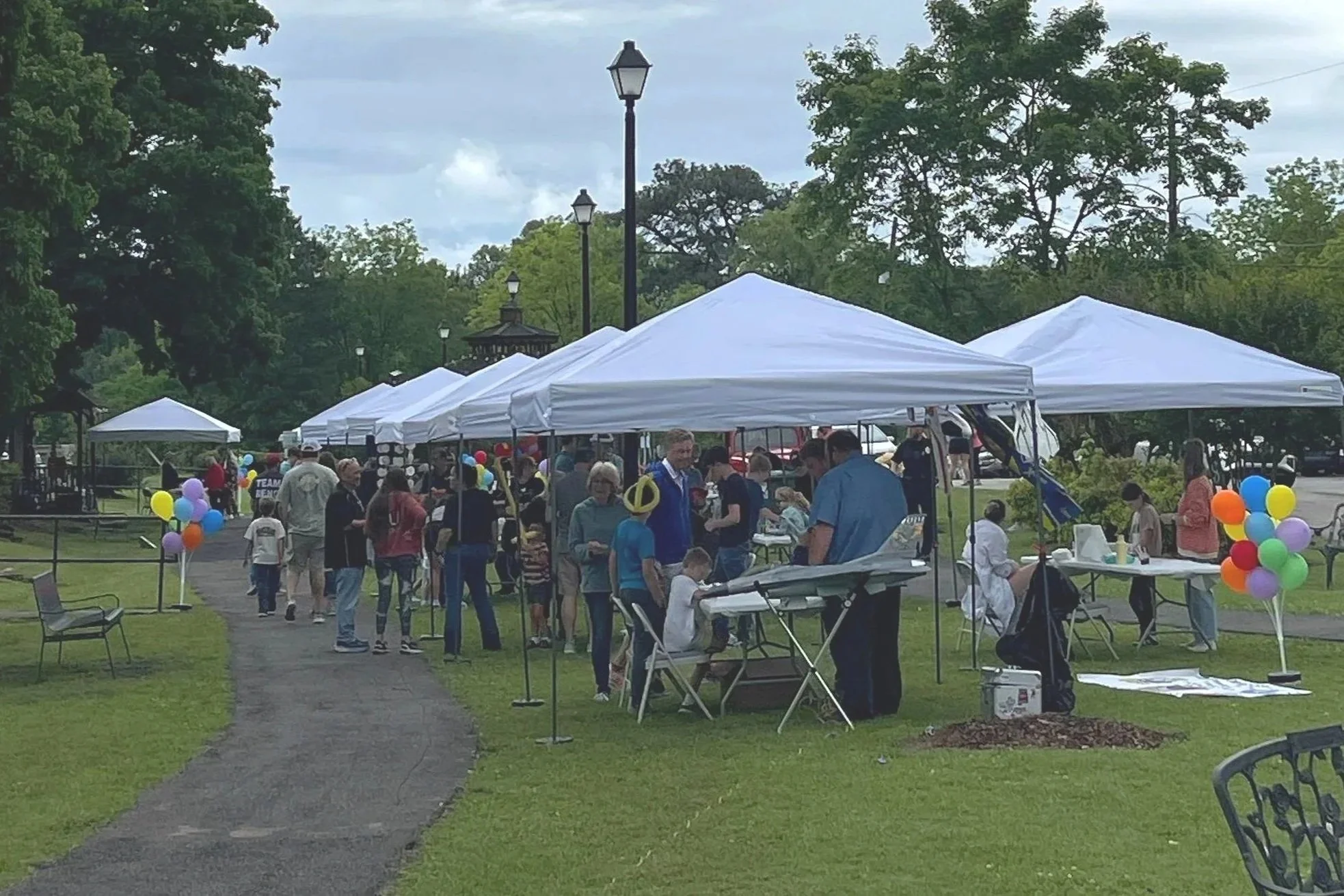 A community outdoor event with people gathered under white canopy tents, balloons, and tables in a park with trees and lampposts in the background.