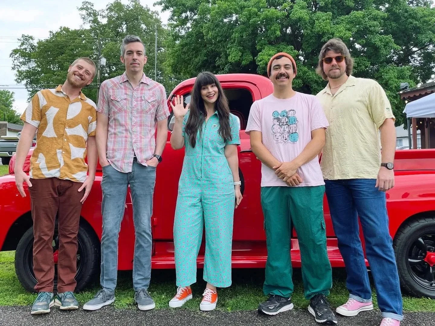 Group of six friends standing in front of a red pickup truck outdoors, smiling and waving, surrounded by green trees.