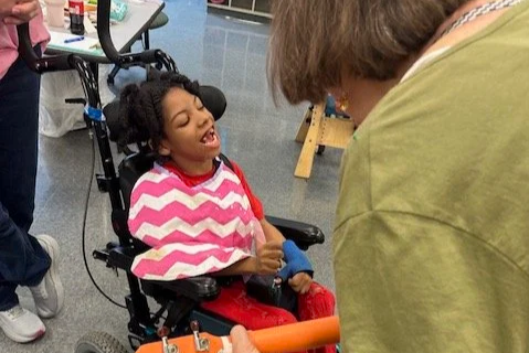 Young girl with a joyful expression in a wheelchair, interacting with an adult.