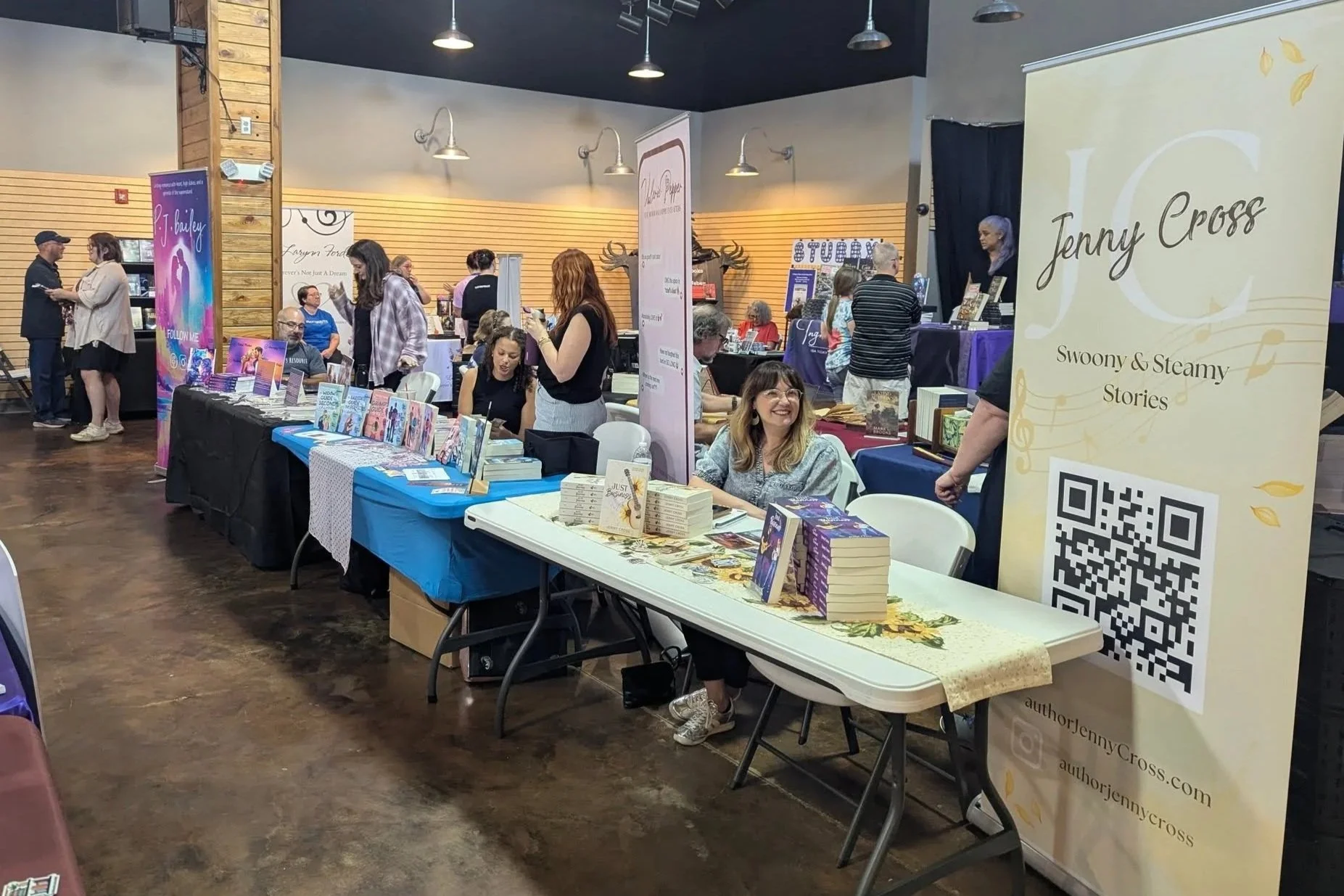 Indoor book fair with multiple tables displaying books, people browsing and chatting, and banners including one for Jenny Cross with a QR code.