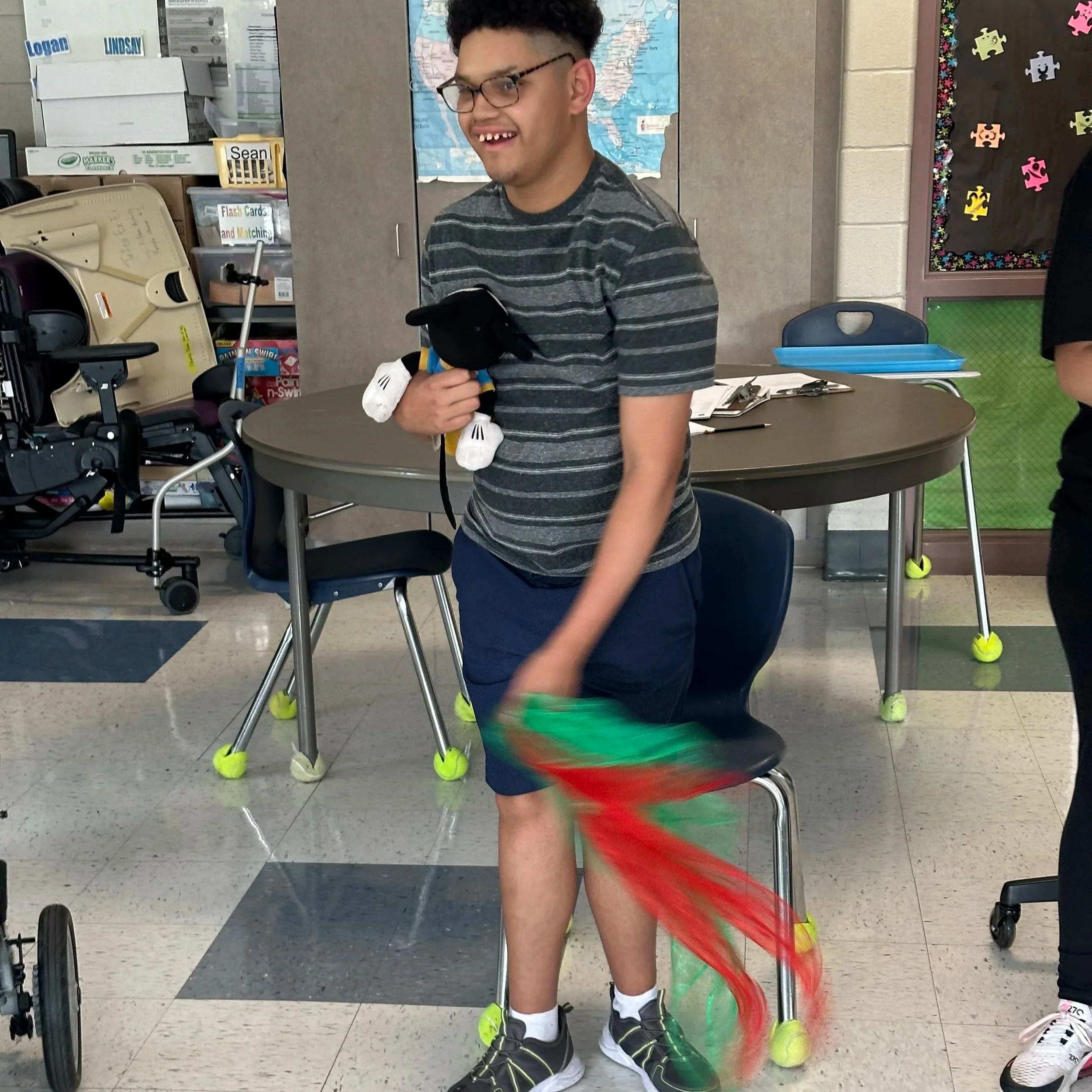 A young man with glasses, dark curly hair, and missing front teeth is standing indoors at a table, holding his hands together. He is wearing a gray and black striped t-shirt and dark shorts. Around him are a wheelchair, a green plastic cup, a shoelace on the floor, and some shoes. In the background, there are storage shelves, a map on the wall, and a decorated bulletin board.
