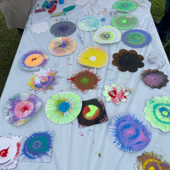Paper plates with colorful, abstract designs created with paint, arranged on a white table covered with paint splatters.