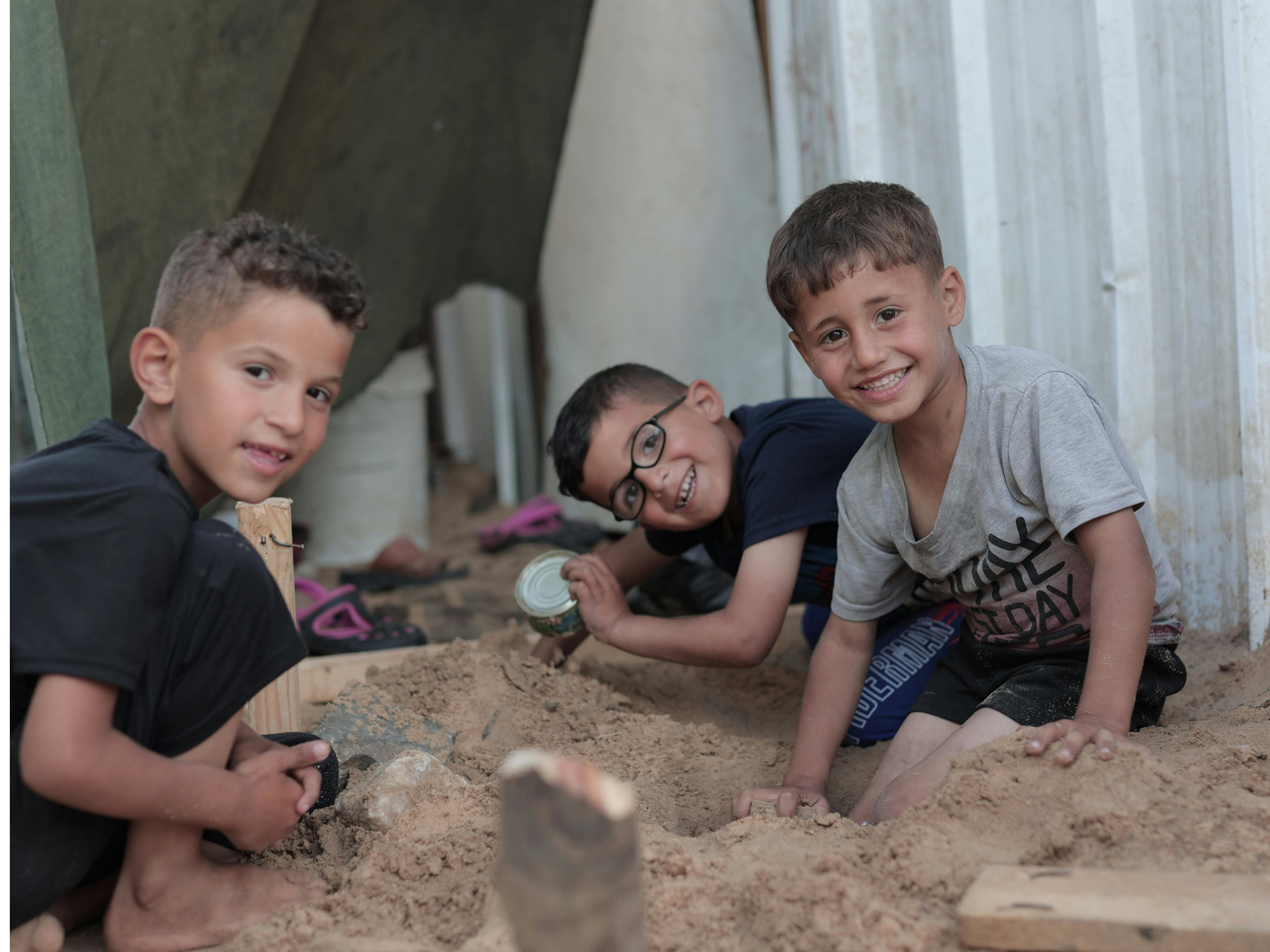 Four boys are sitting in the sand, smiling at the camera. They are under a makeshift shelter made from fabric or tarpaulin. One boy is wearing glasses, and they appear to be playing or working together in an outdoor setting.
