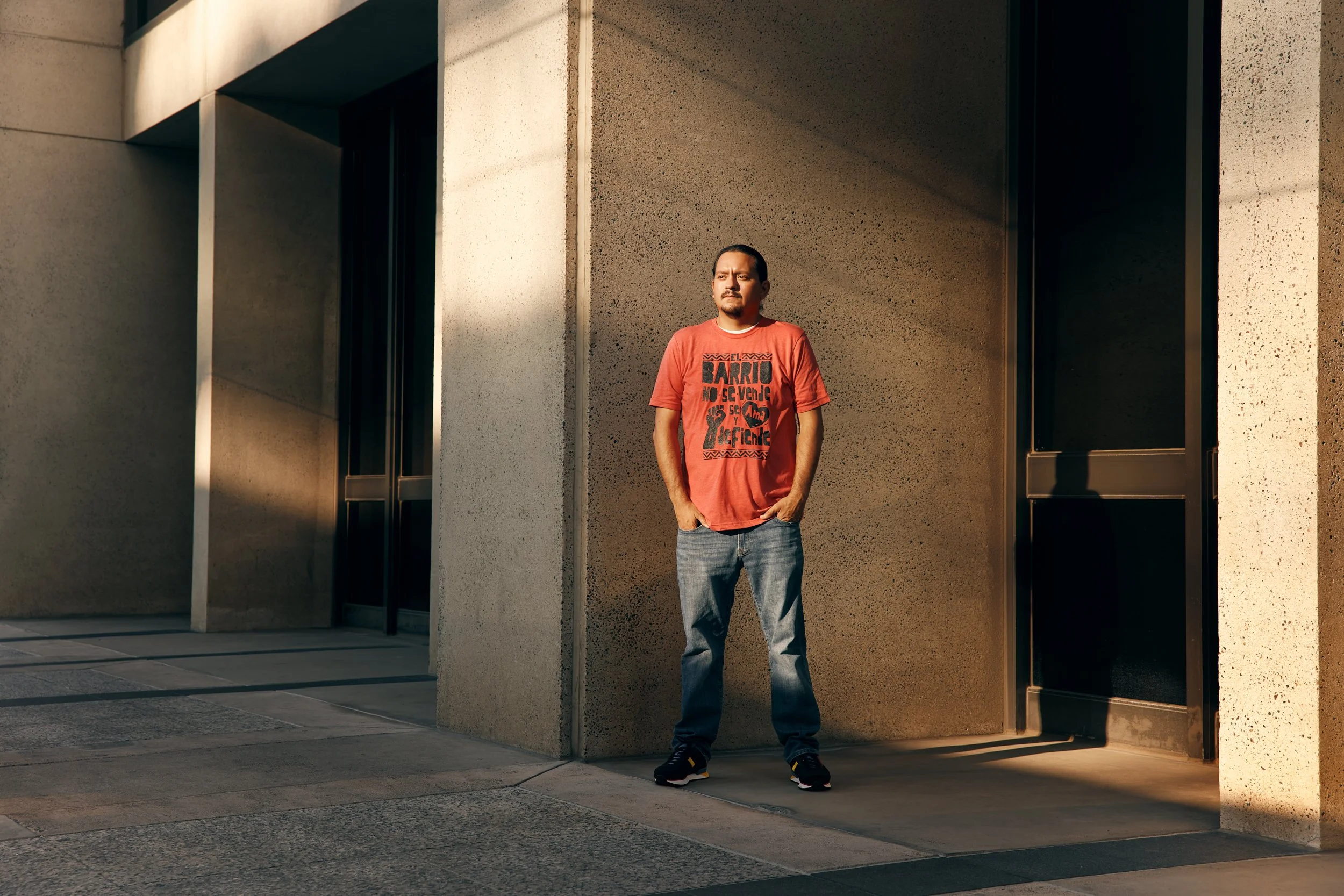 A man with dark hair and a mustache stands outdoors against a large concrete wall, wearing a red graphic t-shirt and jeans, with his hands in his pockets, illuminated by sunlight.