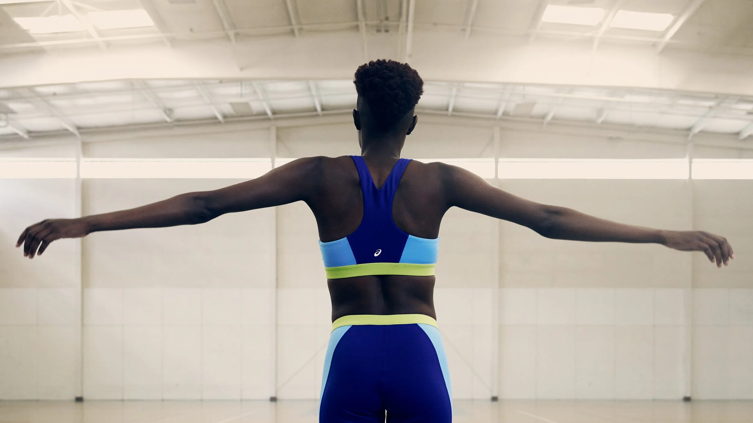 Back view of a female athlete in a blue and yellow sports outfit, standing in a spacious indoor sports hall with arms extended sideways.