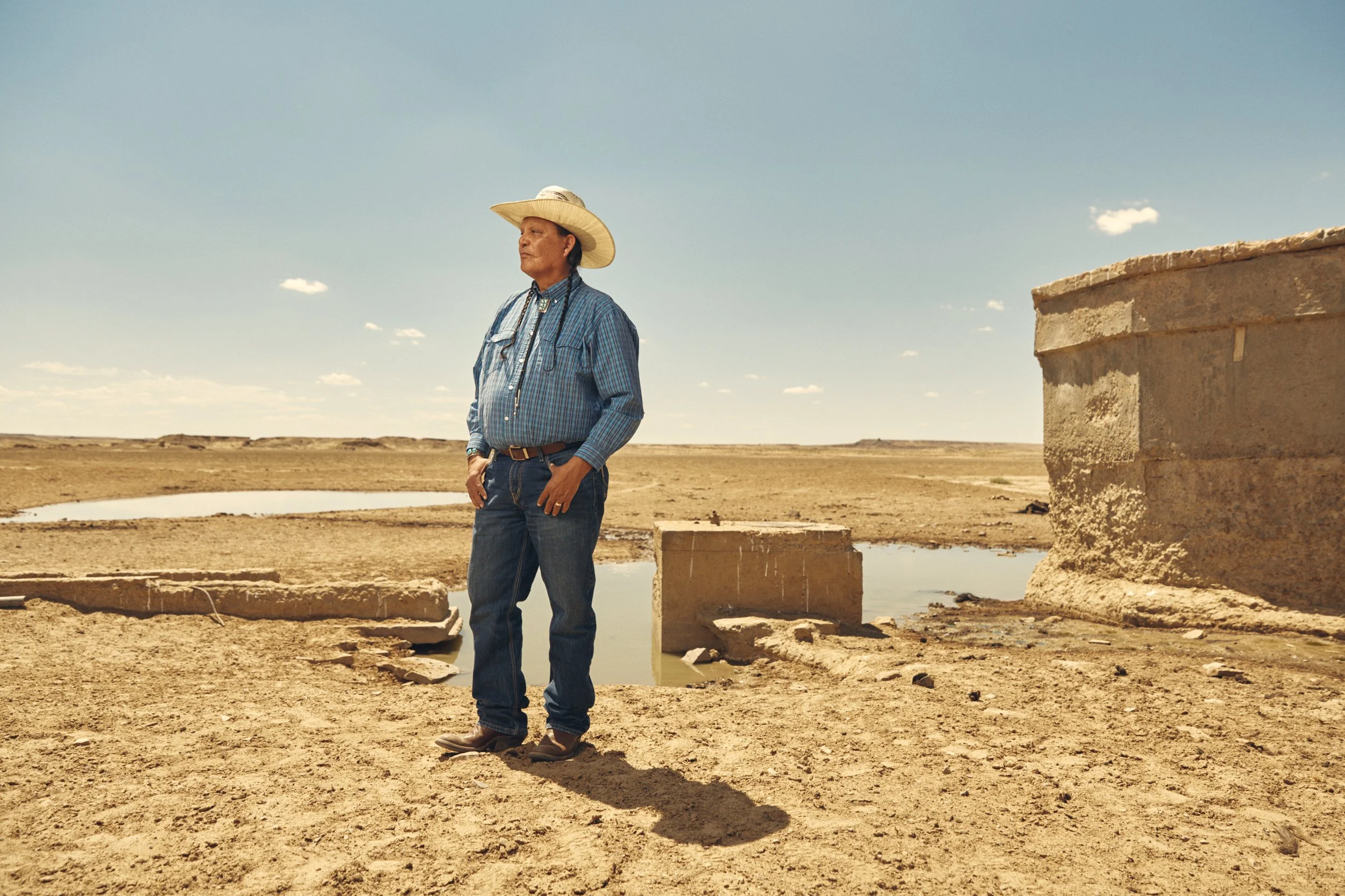 A man wearing a cowboy hat, plaid shirt, and jeans stands outdoors on a barren landscape with a partially broken structure and small pools of water, under a blue sky with scattered clouds.