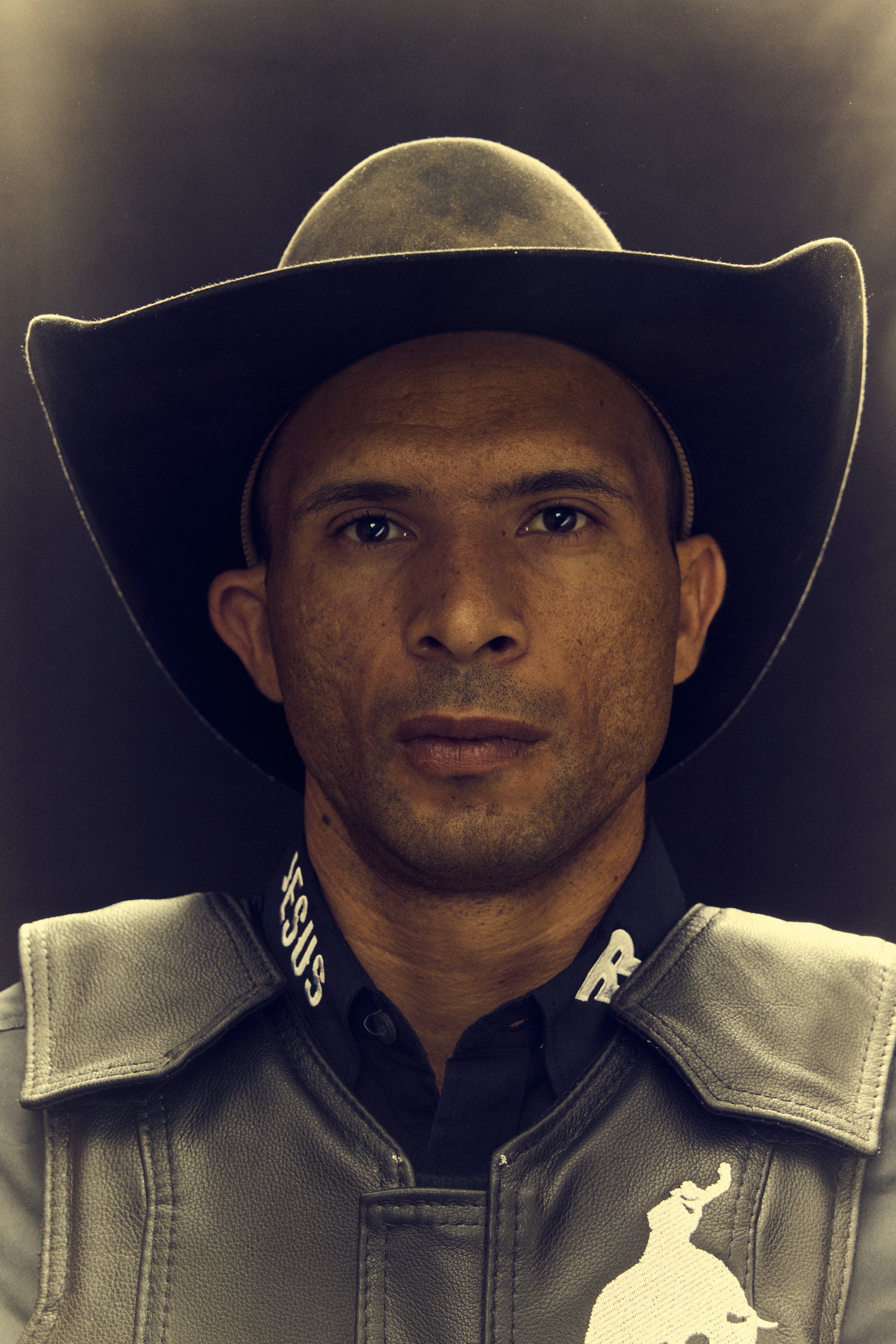 A young man wearing a wide-brimmed hat and a leather jacket, with a serious expression, indoor setting with dark background.